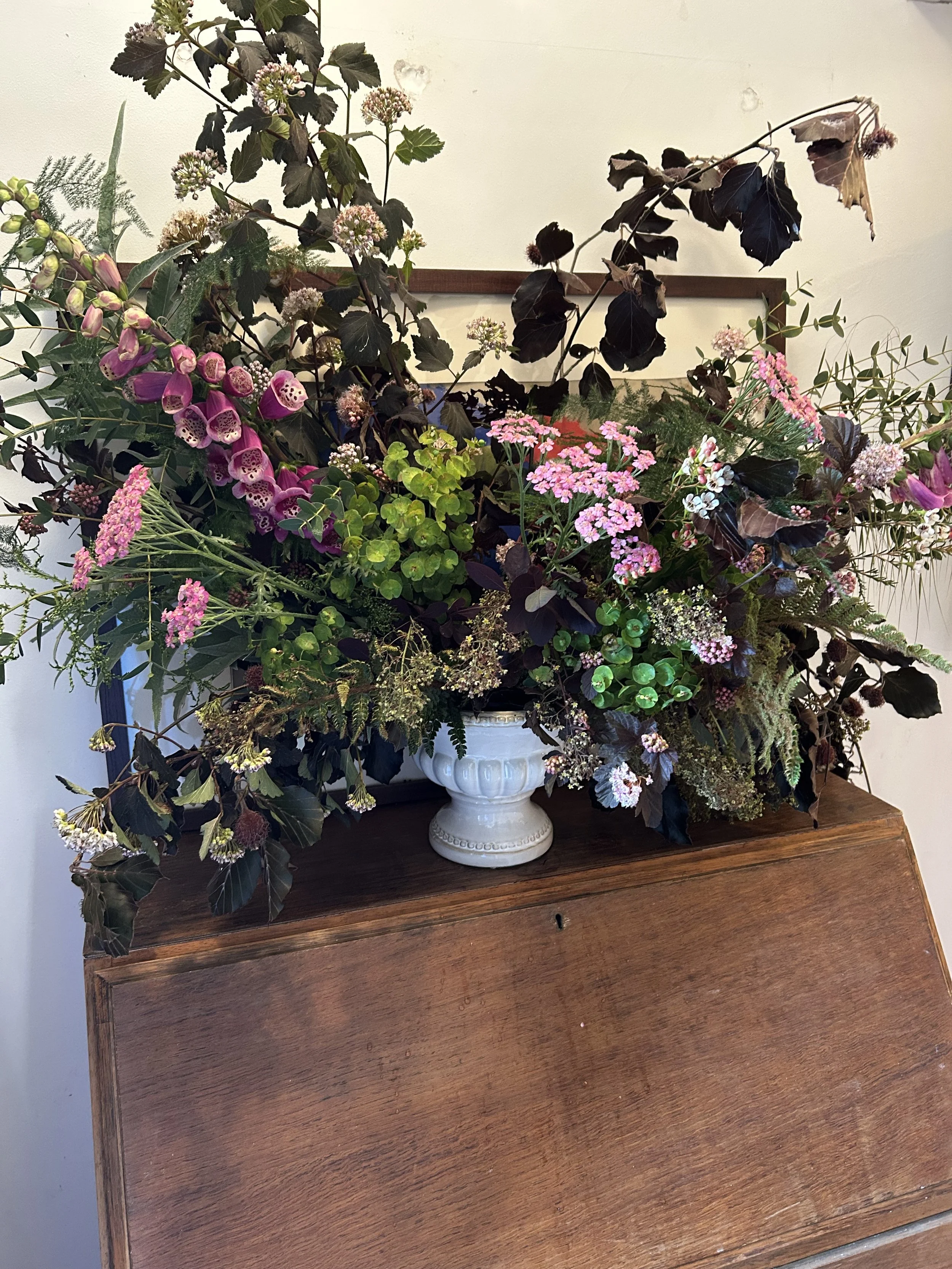 A large floral arrangement in a white vase placed on a wooden table. The arrangement includes pink, purple, and white flowers with dark green and dark purple leaves. The background shows a plain white wall with a wooden trim.