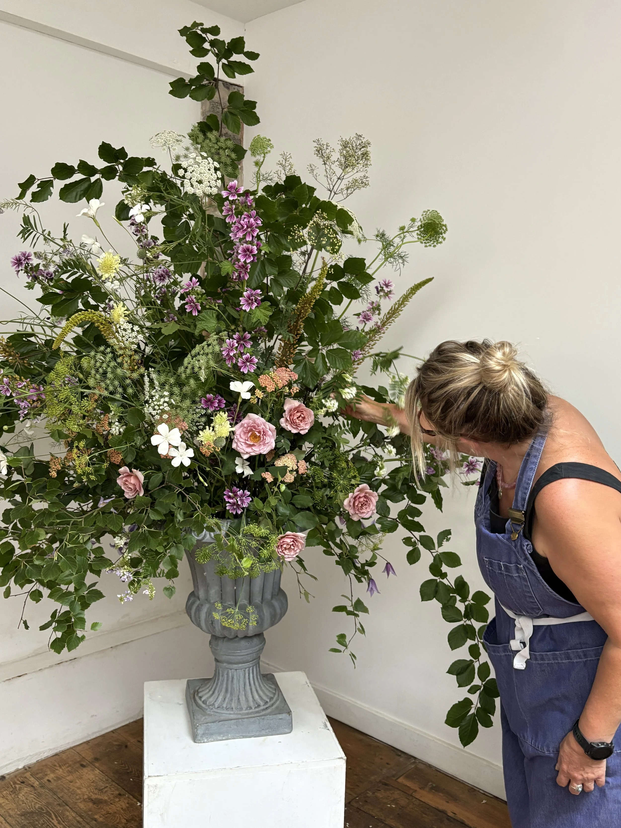 A woman in a denim apron arranging a large floral display on a pedestal in a room with white walls and wooden flooring.