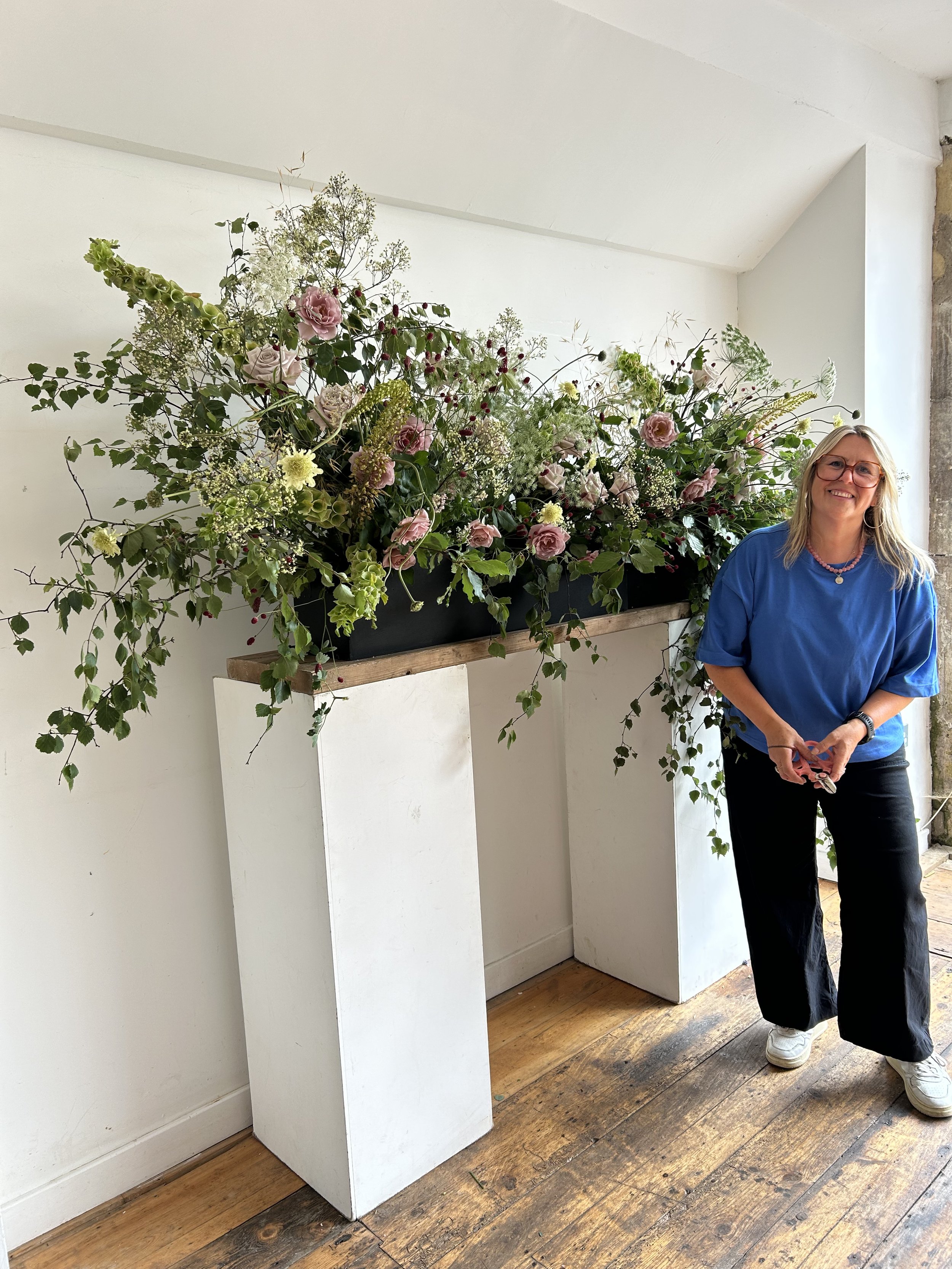 A woman with blonde hair, glasses, and a blue top stands next to a large arrangement of flowers on white pedestals inside a room with wooden floors and white walls.