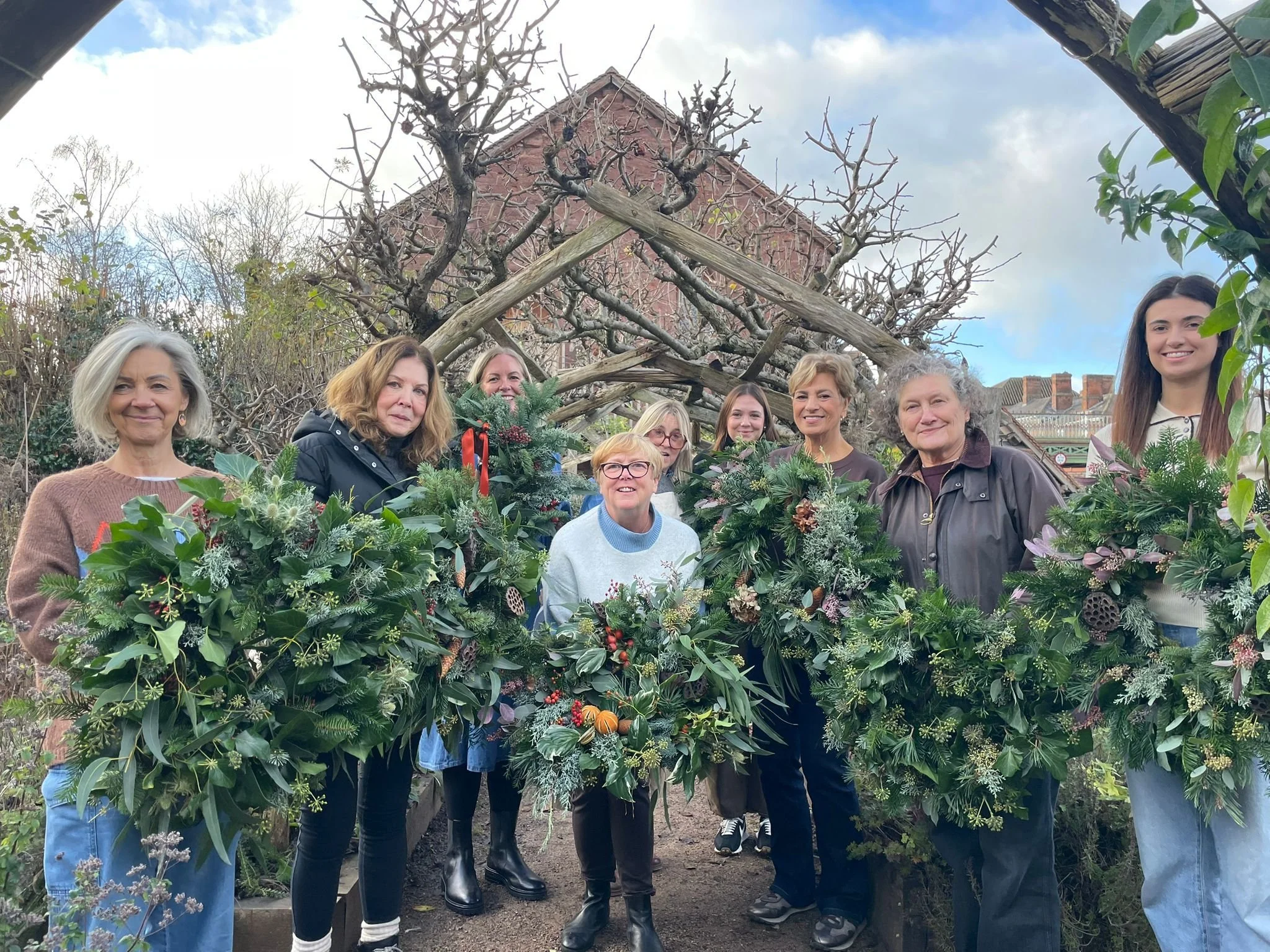 A group of women standing outdoors behind a holiday wreath-covered archway, with a leafless tree and house rooftops in the background.