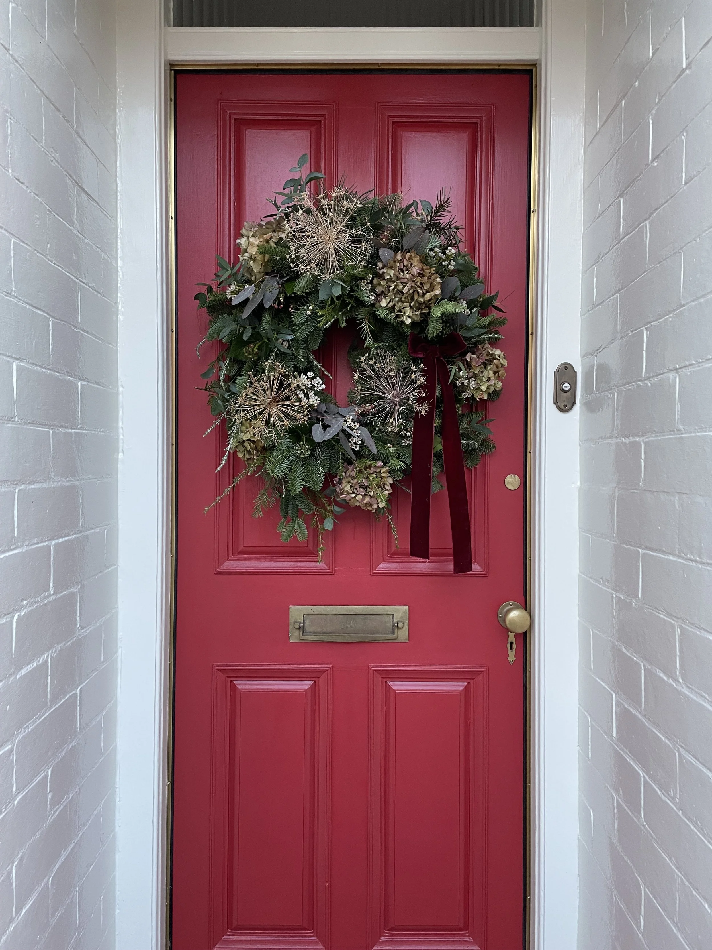 A red front door decorated with a wreath made of greenery, dried flowers, and a burgundy ribbon, set in a white siding entryway.