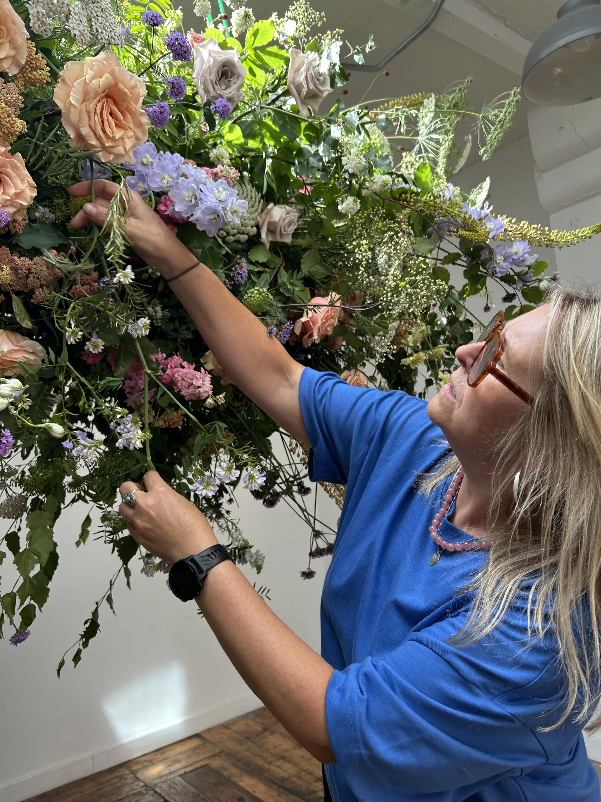 A woman with blonde hair, glasses, and a blue shirt is arranging a large bouquet of colorful flowers. She is wearing a beaded necklace and a watch, and is pulling a flower with her left hand while touching the bouquet with her right hand. The backgro