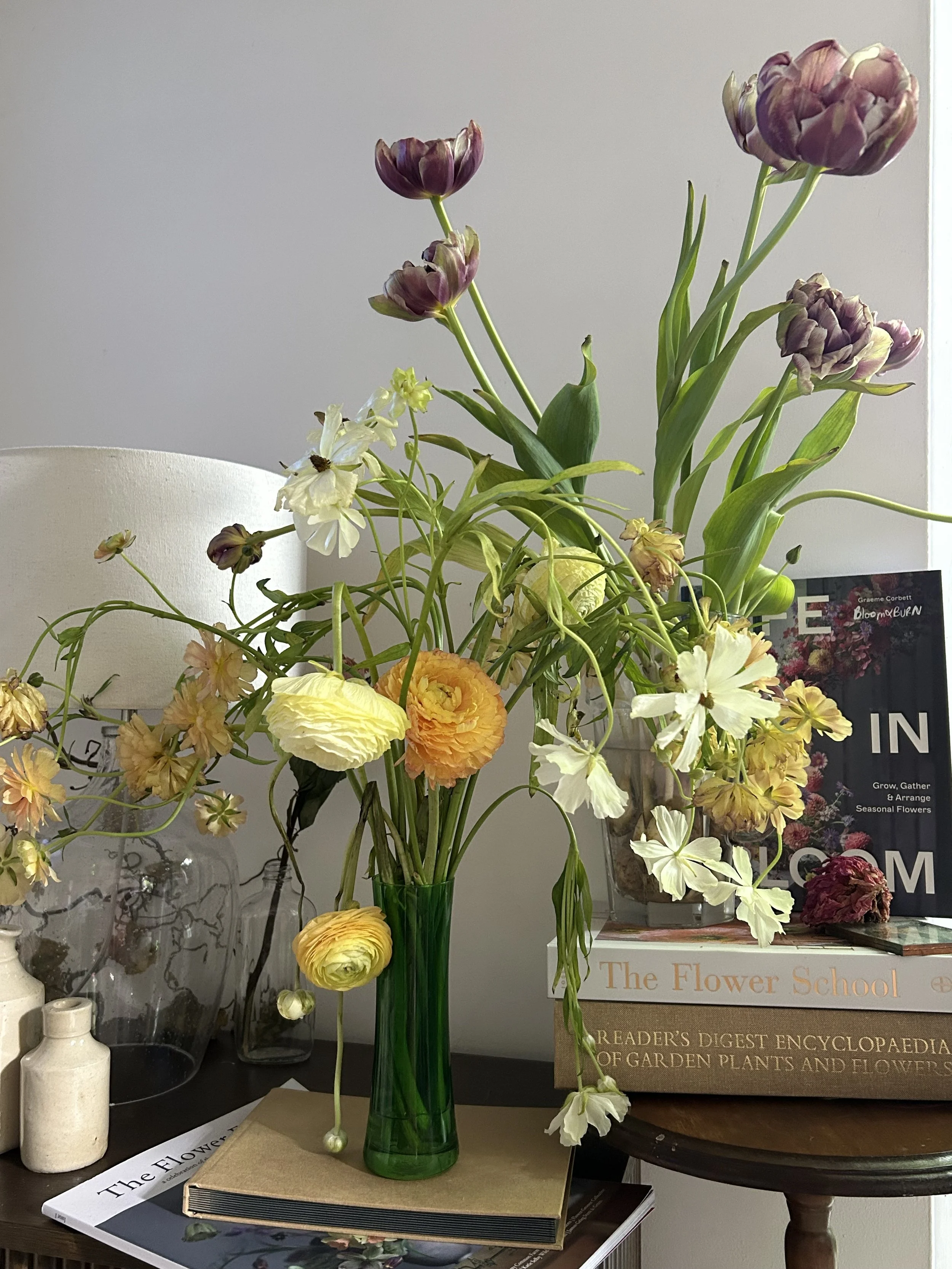 A green glass vase with an arrangement of various flowers, including purple, white, and yellow blossoms, placed on a table with books and decorative items in the background.
