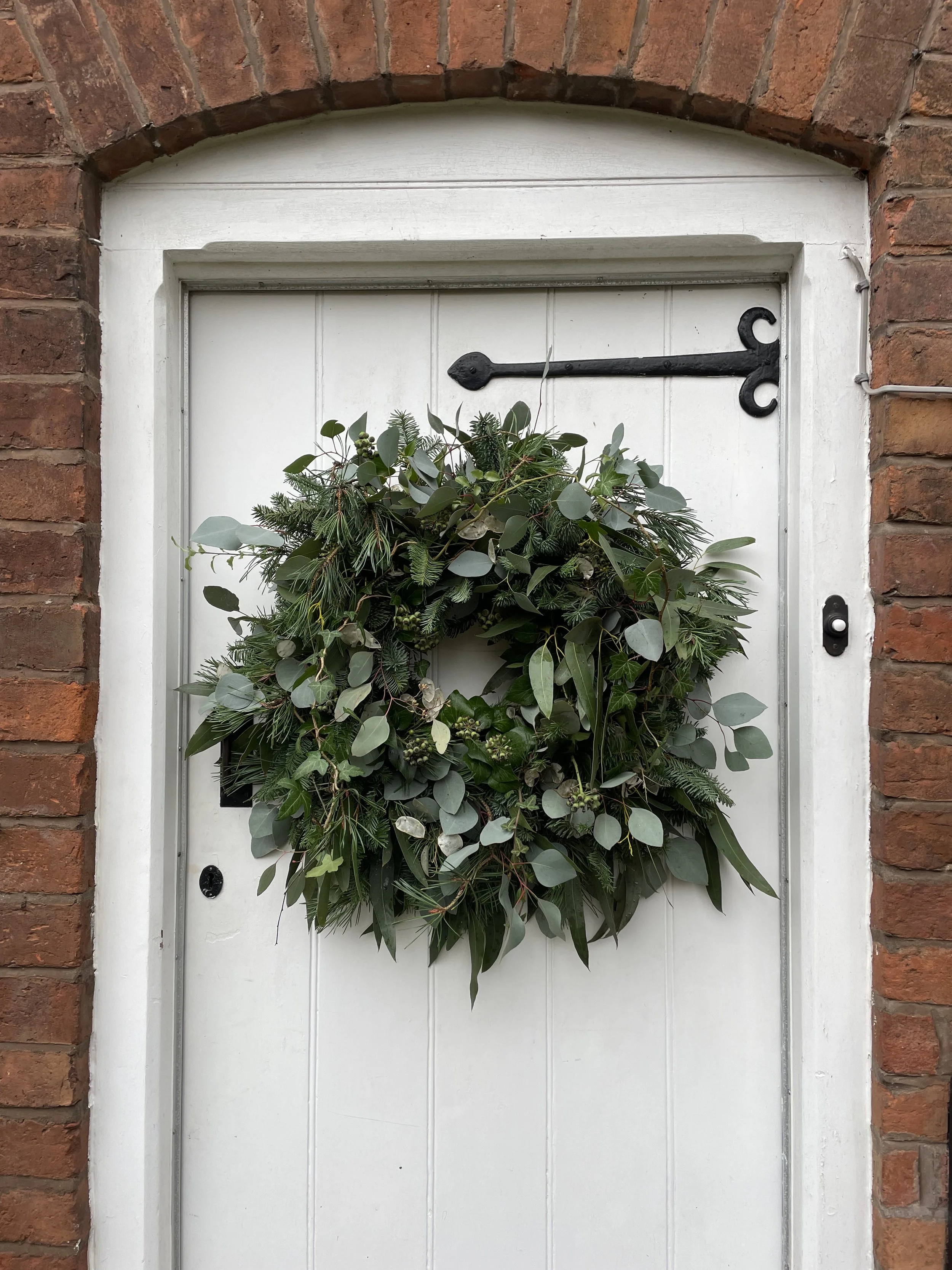 A white door with a black decorative hinge, surrounded by a brick wall. A green wreath made of various leaves is hanging on the door.