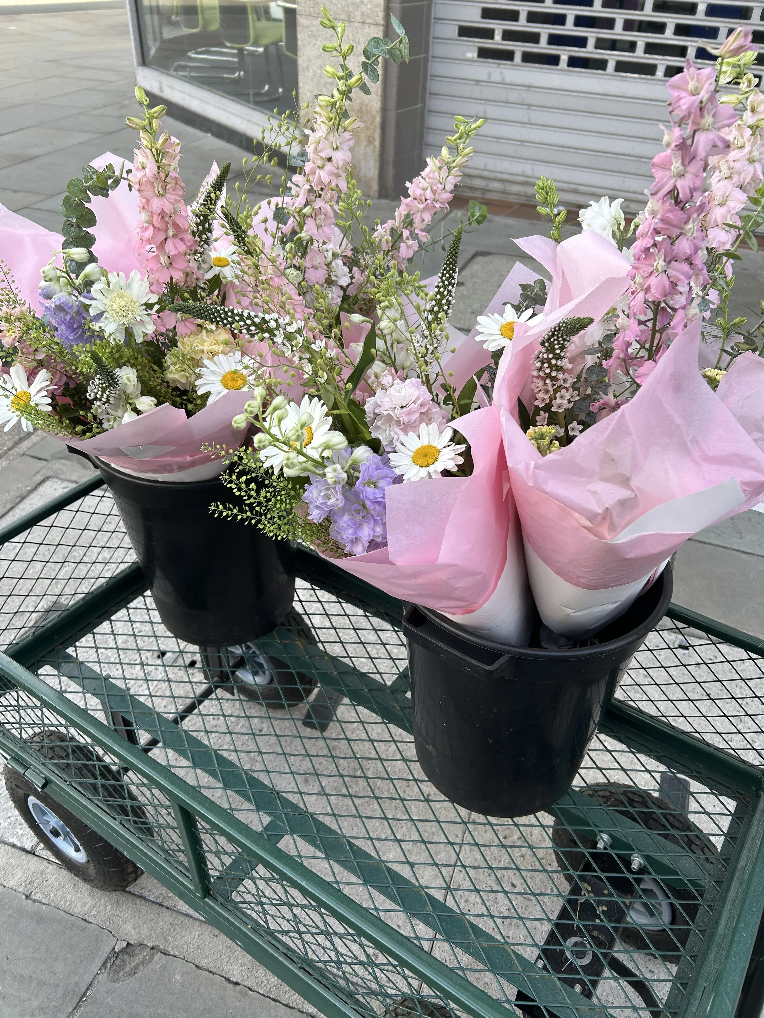 A shopping cart with two black flower pots containing bouquets of pink, white, purple, and green flowers wrapped in pink tissue paper, placed on a sidewalk near a store entrance.