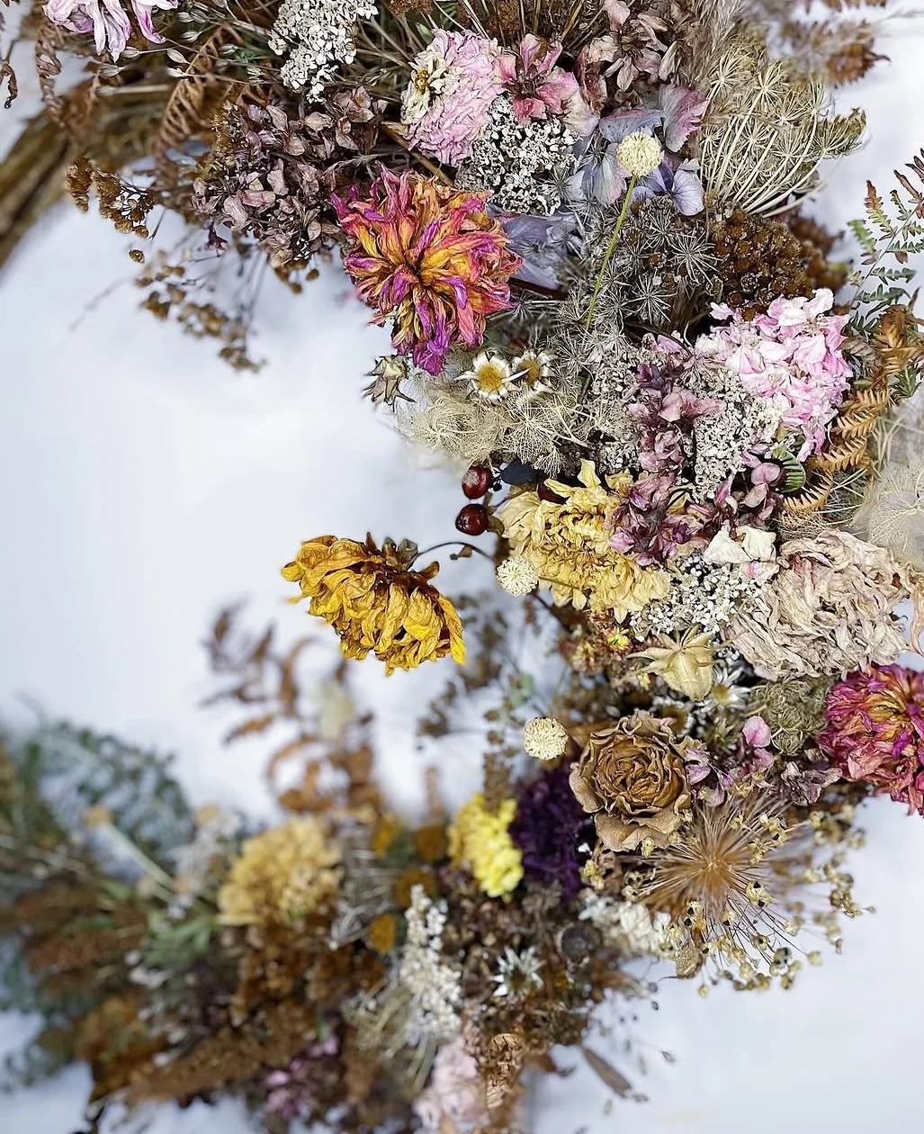 An overhead view of a dried floral wreath consisting of various dried flowers and plants in muted colors, including pink, purple, yellow, brown, and white, arranged on a white background.