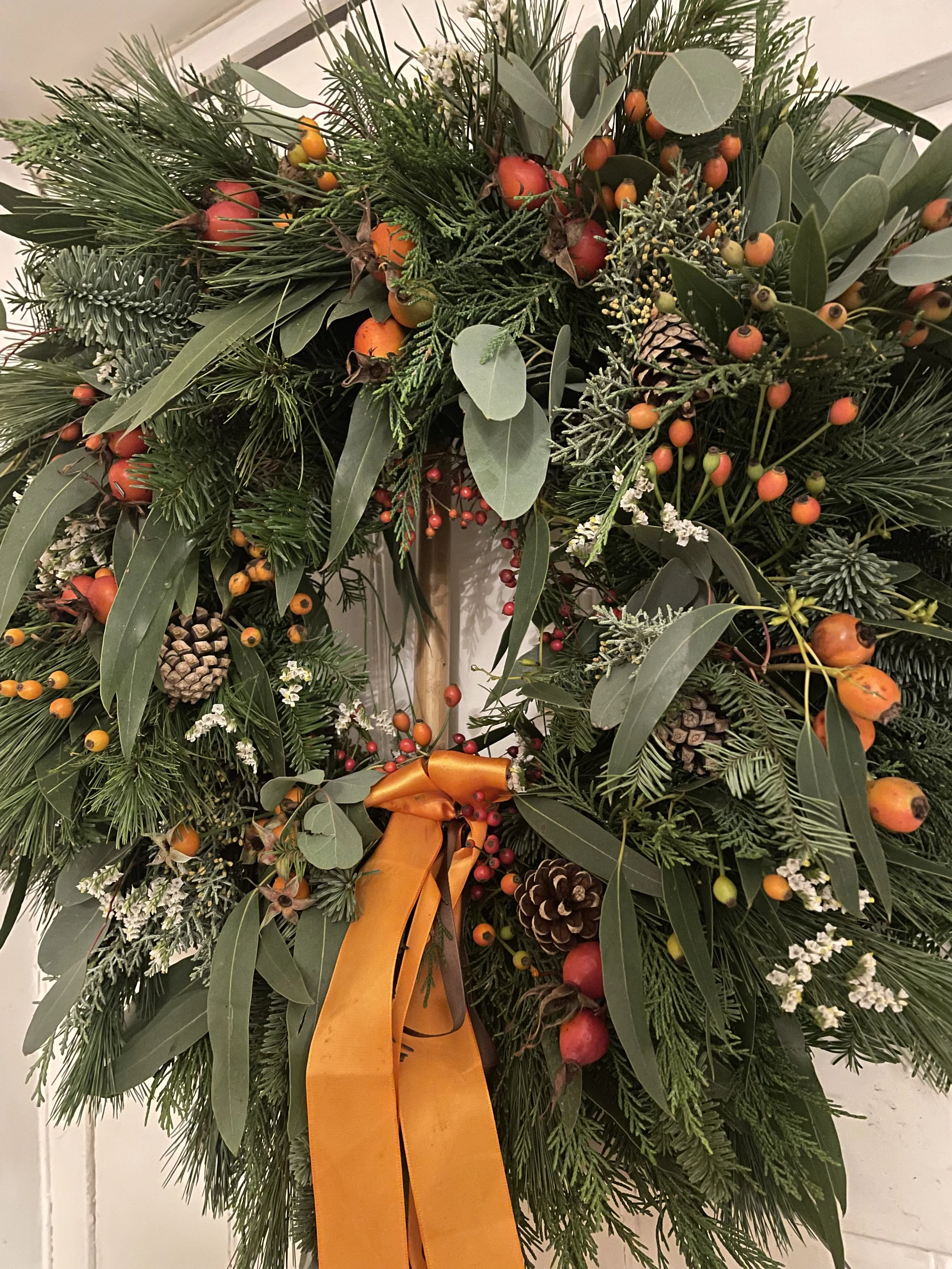 A lush Christmas wreath featuring various evergreen branches, pinecones, small orange and red berries, and a yellow ribbon in the center, hanging on a wall.