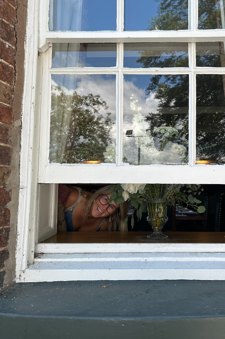 A woman with glasses leaning through a lower window opening, smiling, with a large floral arrangement on the windowsill.