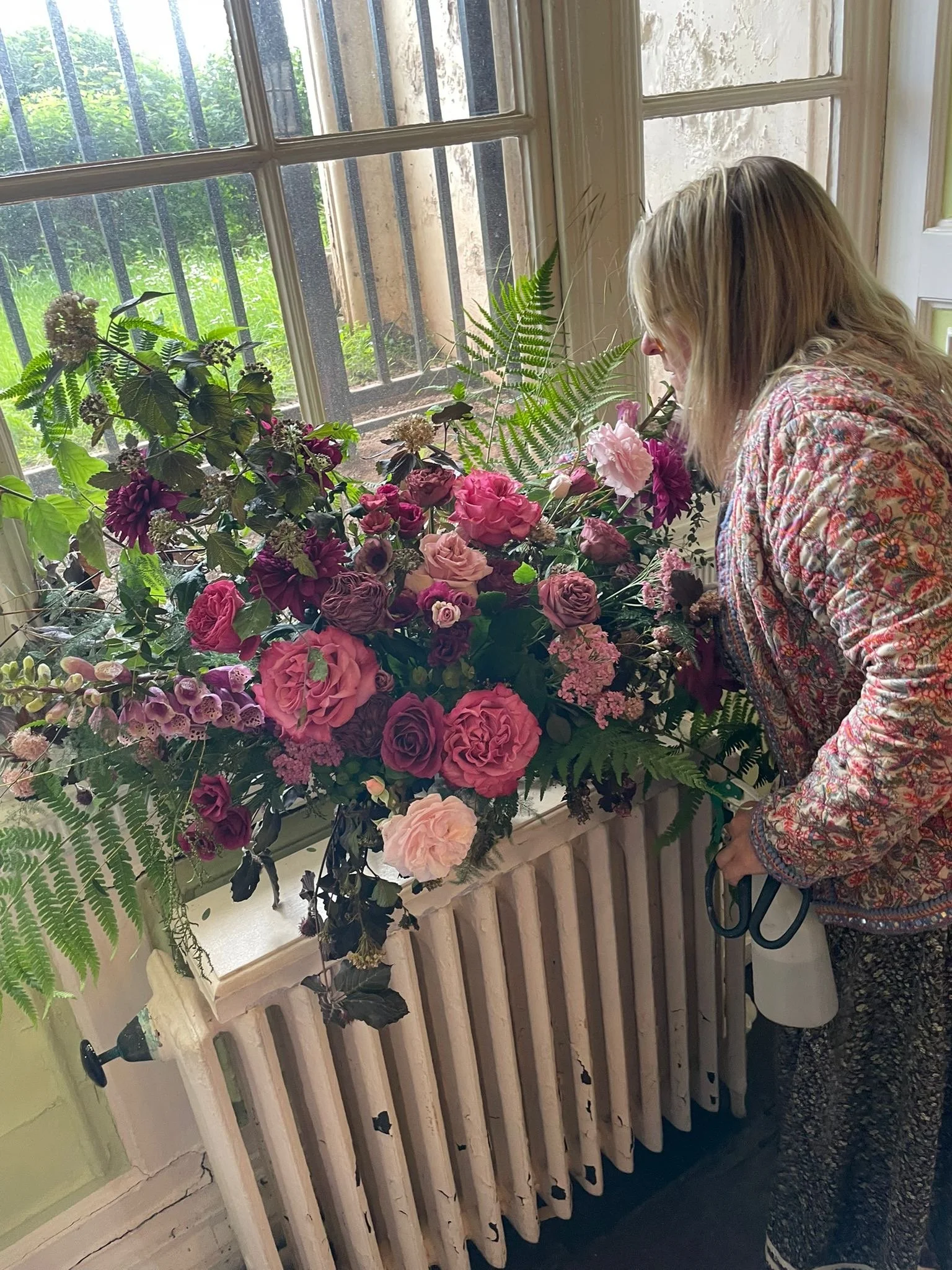 A woman with blonde hair using scissors to arrange a large bouquet of pink, purple, and white flowers, plants, and ferns on a windowsill in a room with a white wooden window frame and a green wall.