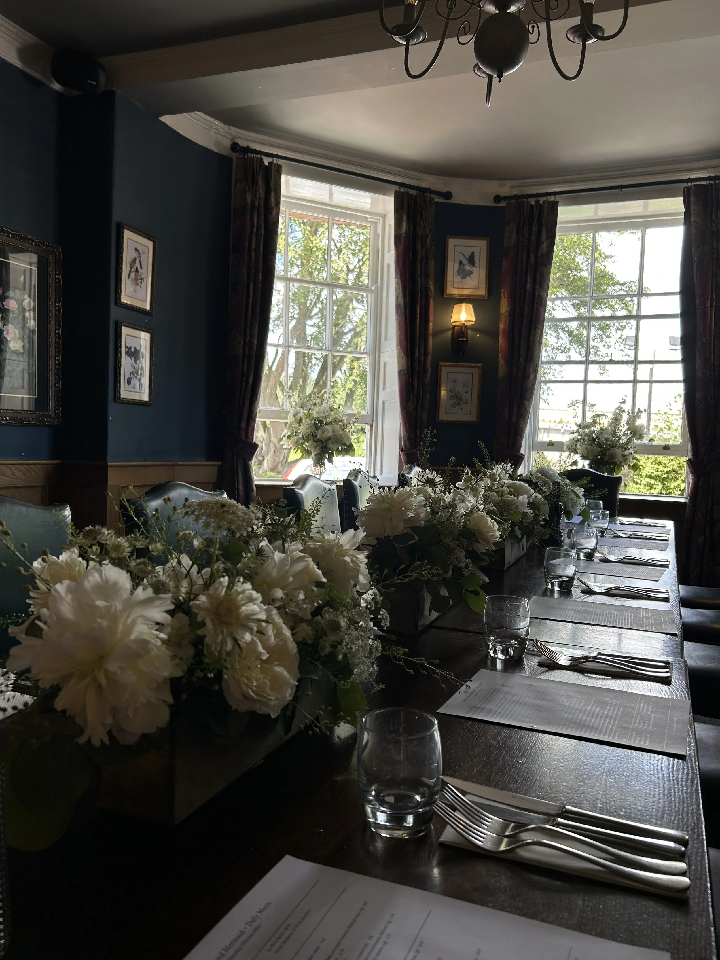 A formal dining room with a dark wooden table set with placemats, silverware, and glasses. The table has a large floral centerpiece with white flowers. The room has large windows with greenery outside, dark curtains, framed artwork on teal walls, a c
