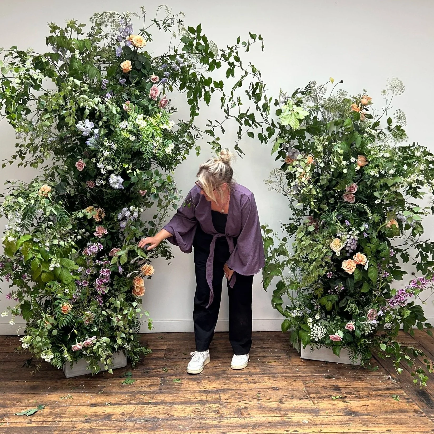 A woman in a purple jacket, black pants, white sneakers, and glasses is standing in front of a white wall and wooden floor, surrounded by tall, lush floral arrangements with various green leaves and blooming flowers in pastel colors.