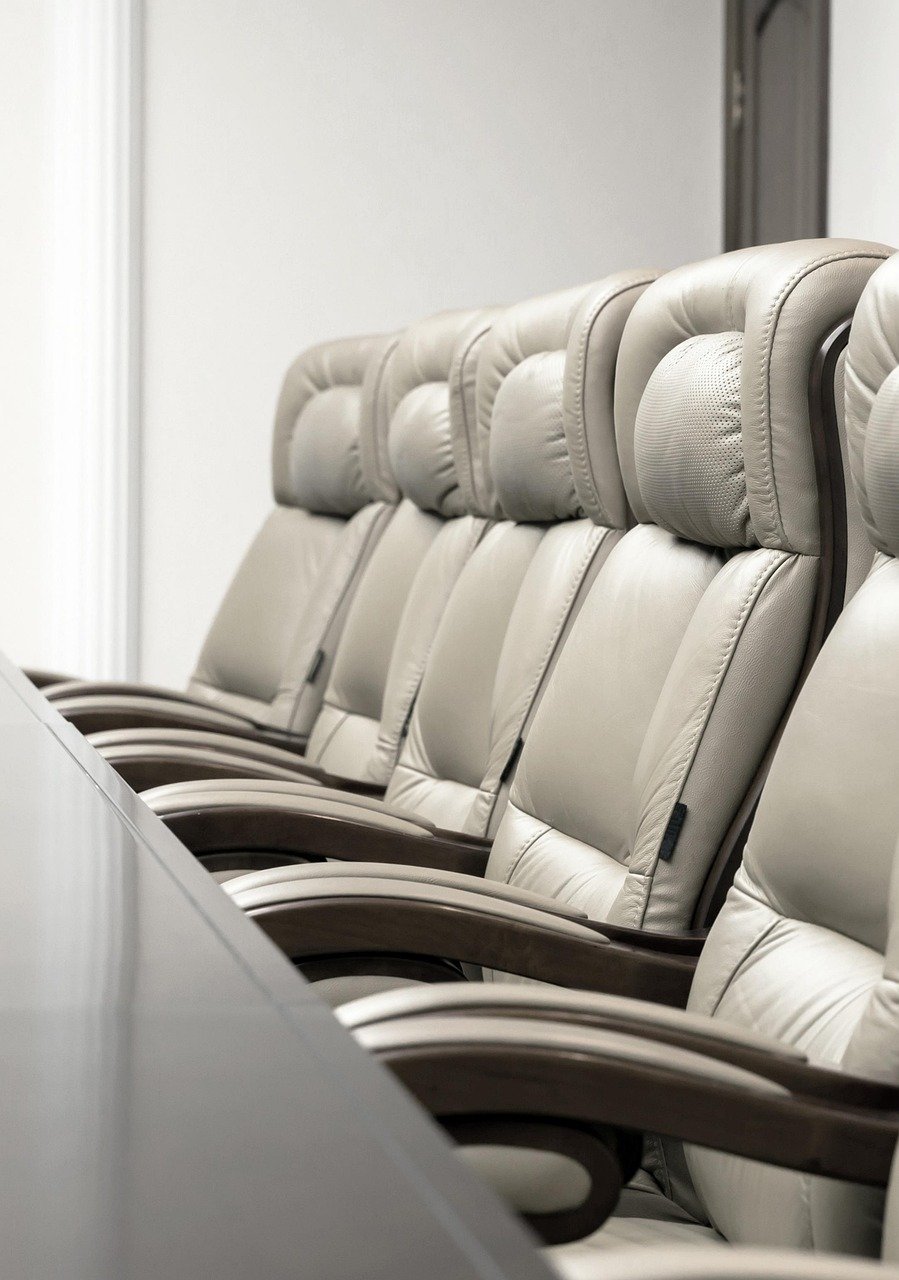 A row of empty, beige leather conference chairs with high backs and armrests, lined up along a table in a meeting room.
