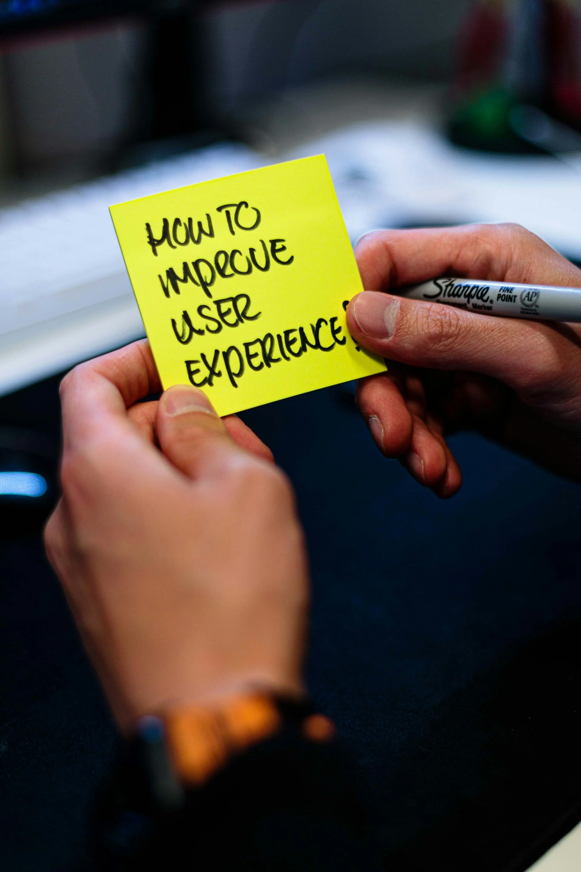 Person writing on a yellow sticky note that reads 'How to improve user experience' with a black Sharpie marker.