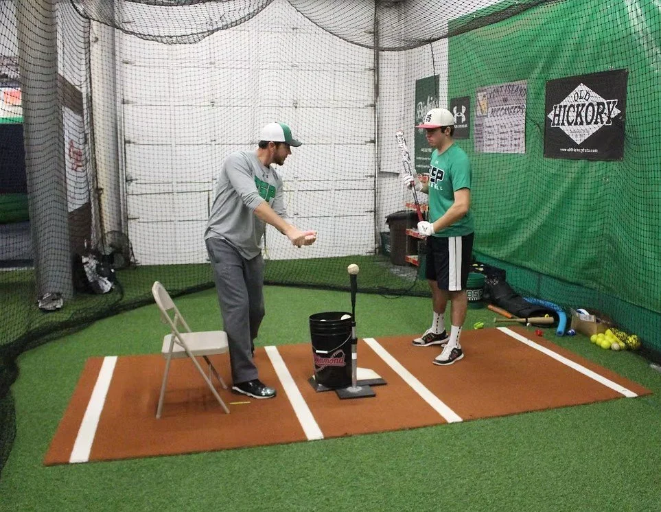 Two men practicing baseball batting inside a batting cage with artificial turf and netting, one man with a bat and the other with a ball, near a bucket of baseballs.