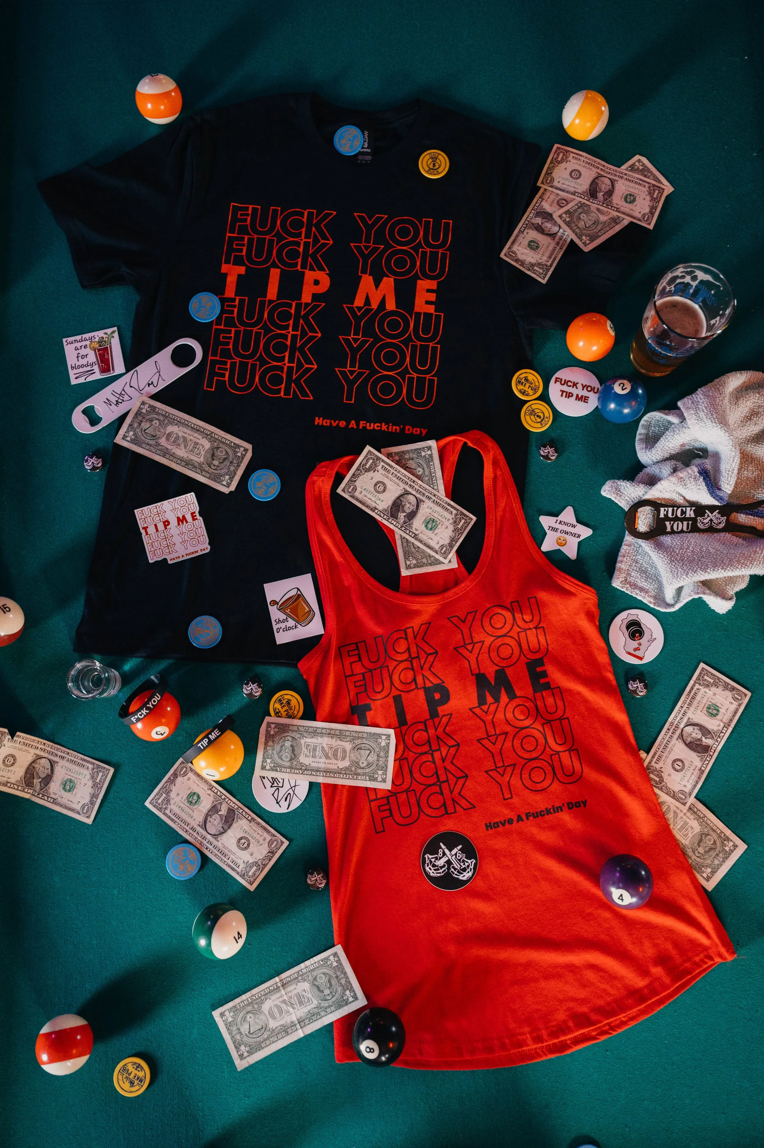 A collection of items on a green surface includes two tank tops with the phrase 'F*** You, Tip Me' and 'Have a F***in' Day' printed in bold red and black letters, dollar bills, assorted Billiard balls, various stickers and buttons with slogans, a glass of beer, a white towel, and some signed memorabilia.