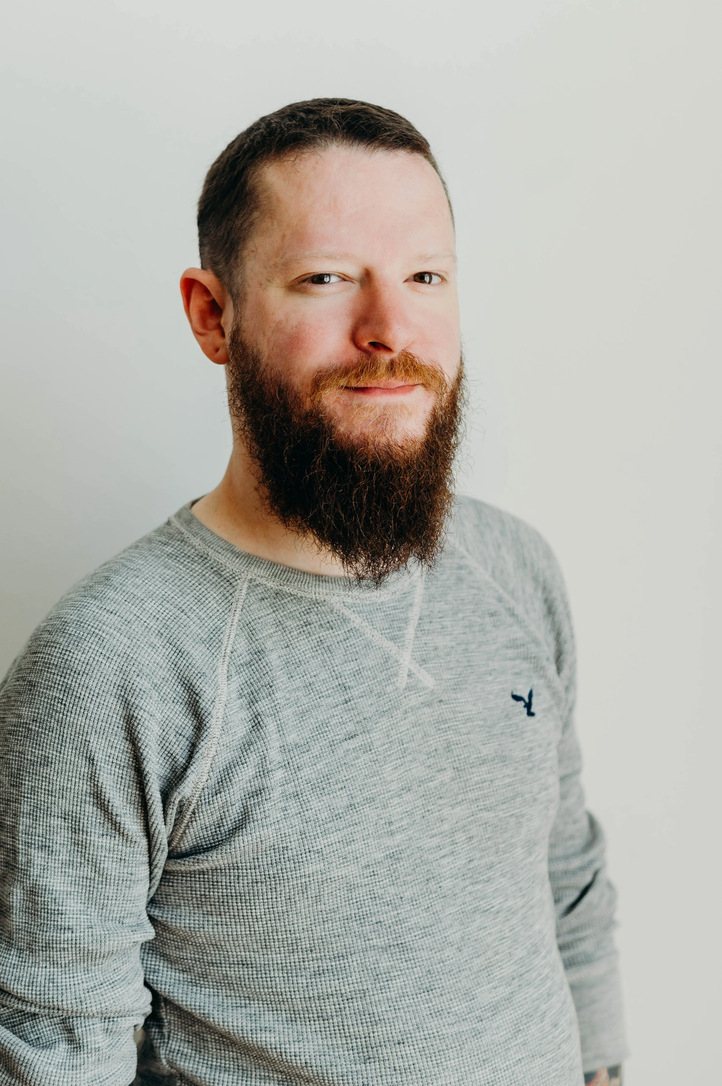 A man with short dark hair, a beard, and light skin wearing a gray sweatshirt with a small blue logo on the chest, standing against a plain white background.