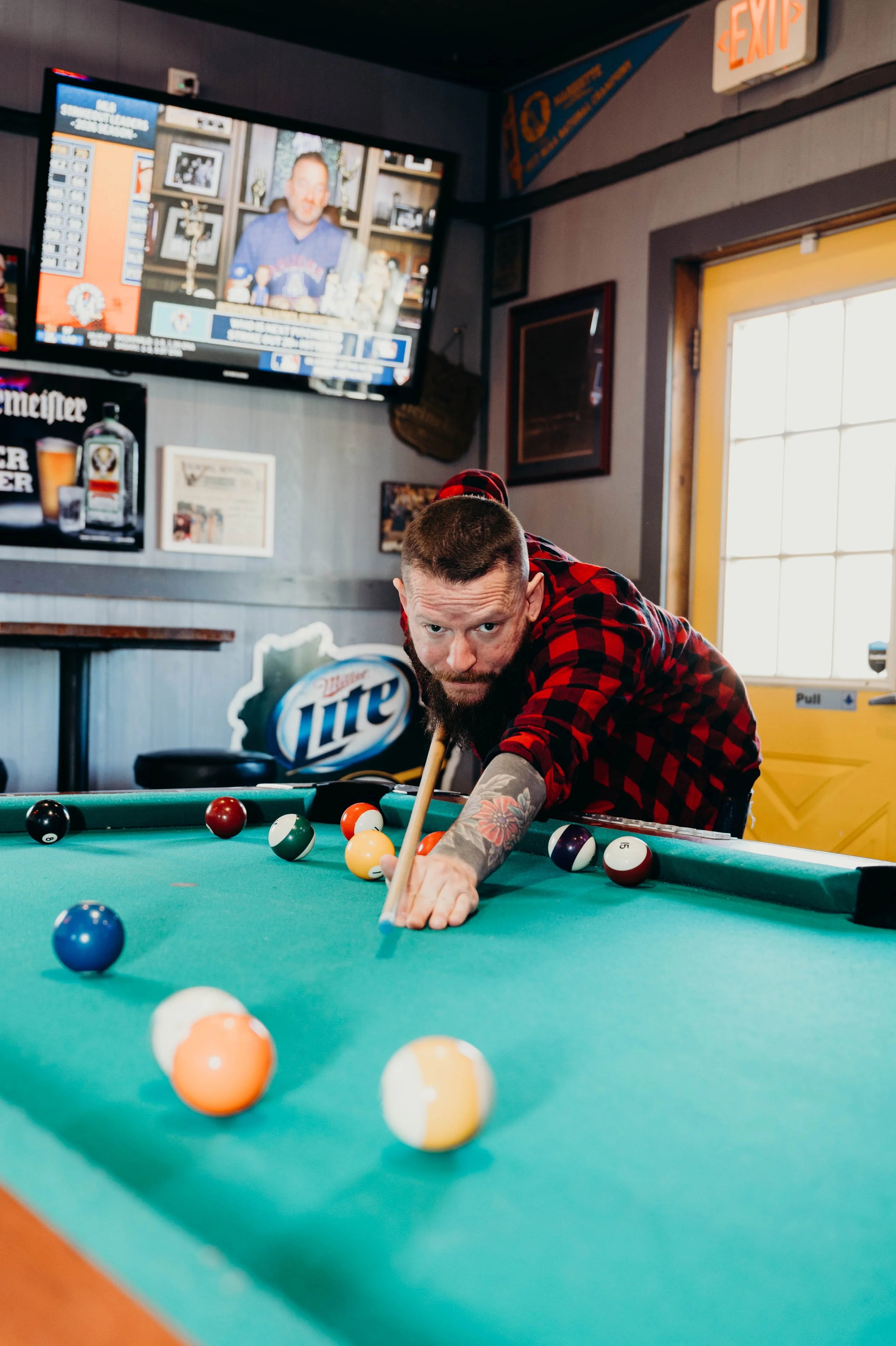 Man with tattoos in a red plaid shirt playing pool. TV screen shows a commentator. Bar with Miller Lite sign, framed pictures, and an exit door are visible in background.