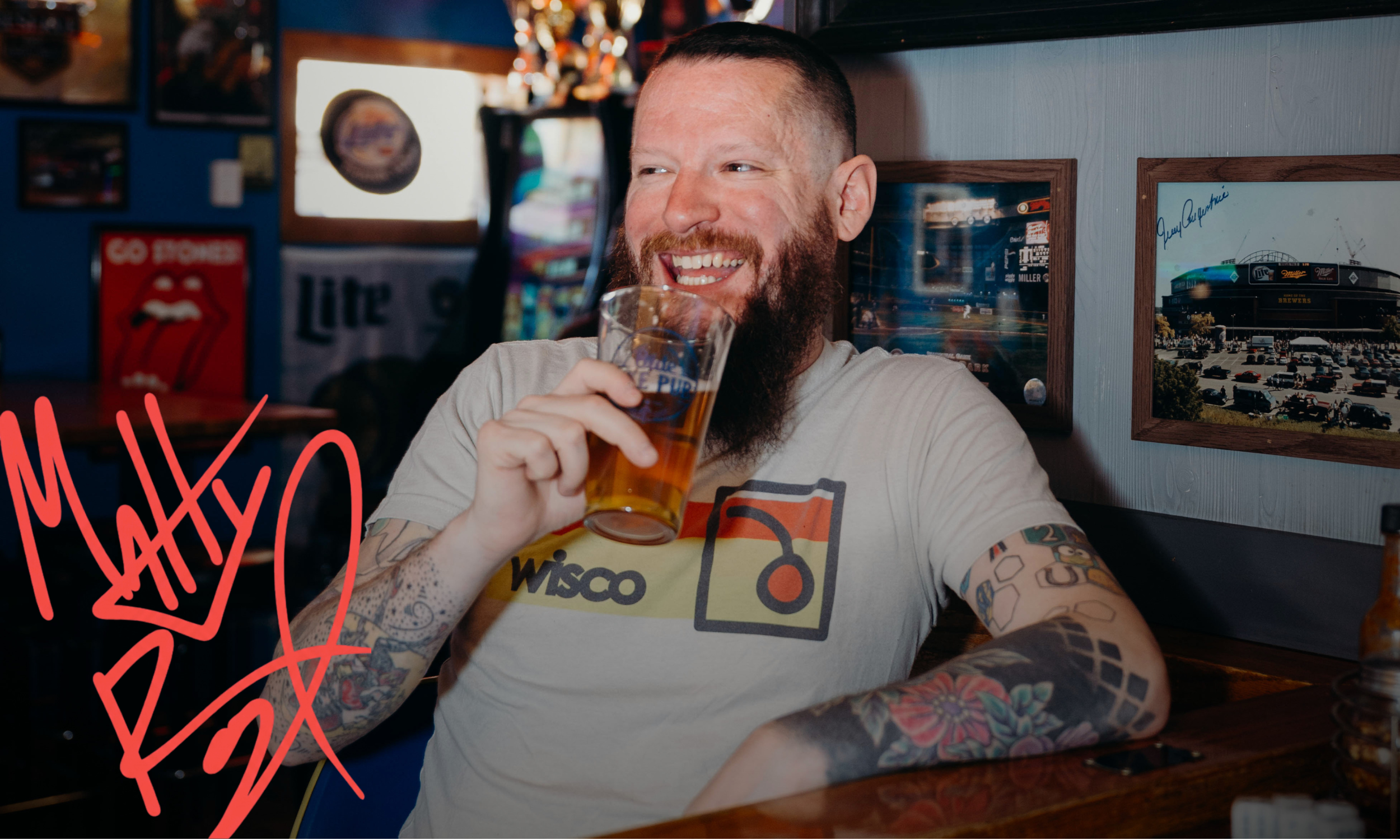 A man with a beard and tattoos on his arms is smiling and holding a glass of beer in a bar or restaurant. The background features framed pictures and posters on the wall, including a Rolling Stones tongue logo, and a large window with natural light.