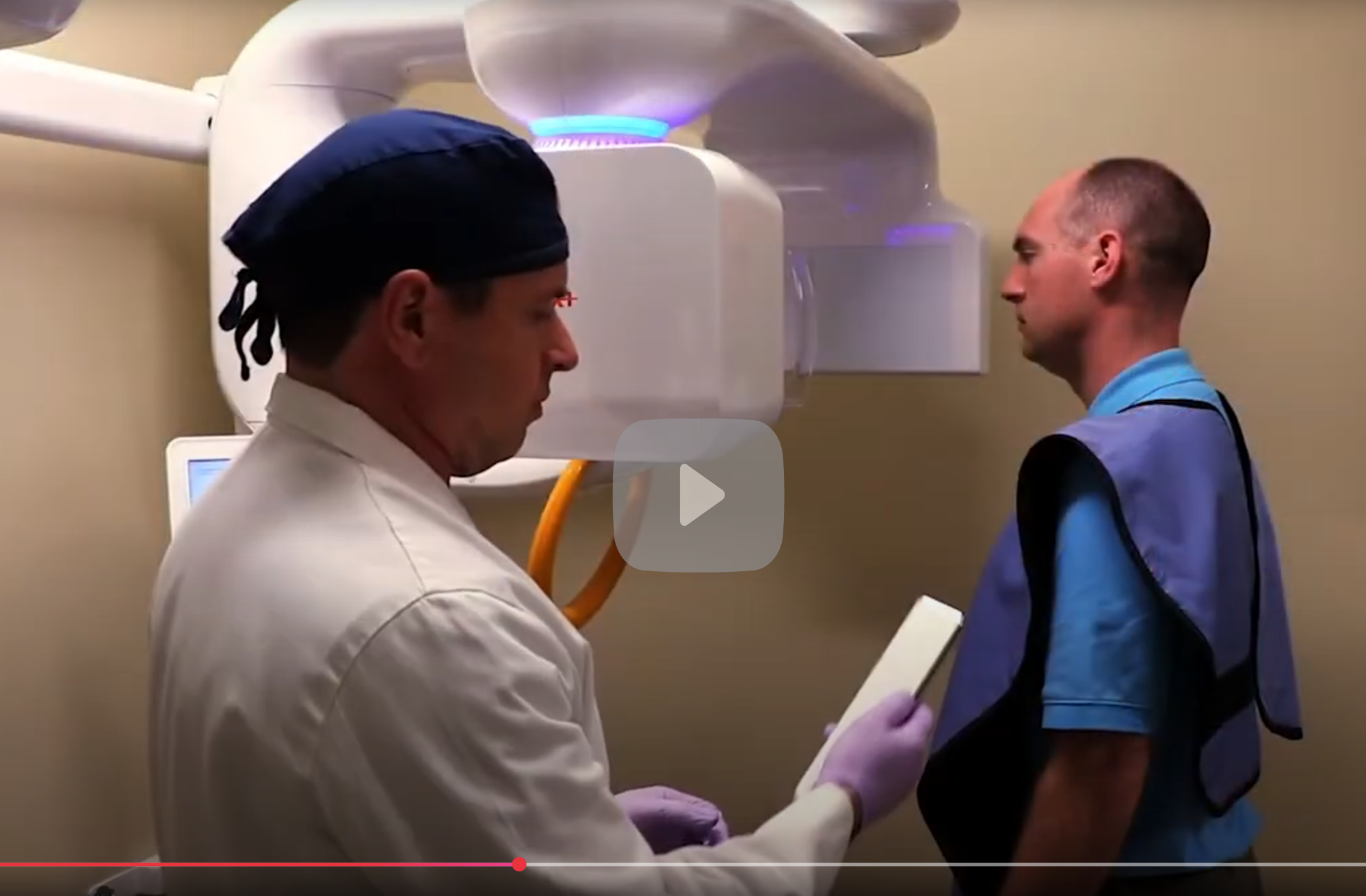 A doctor and a patient in a room with a medical imaging machine. The doctor is looking at a clipboard, and the patient is standing, wearing a lead apron.