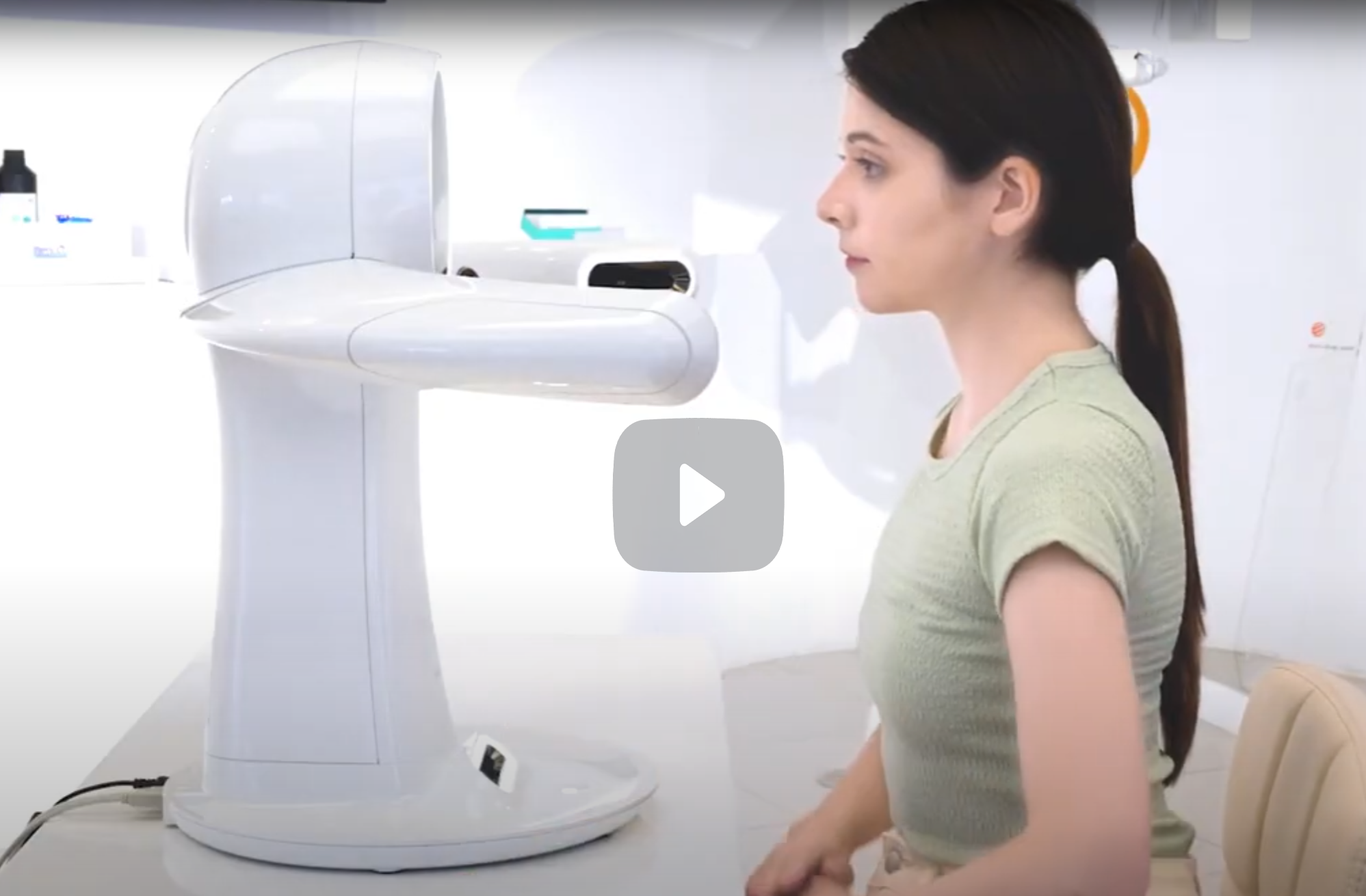 A woman sitting in a hospital or medical clinic, preparing for an imaging scan with a large, white medical device that looks like an MRI or CT scanner.