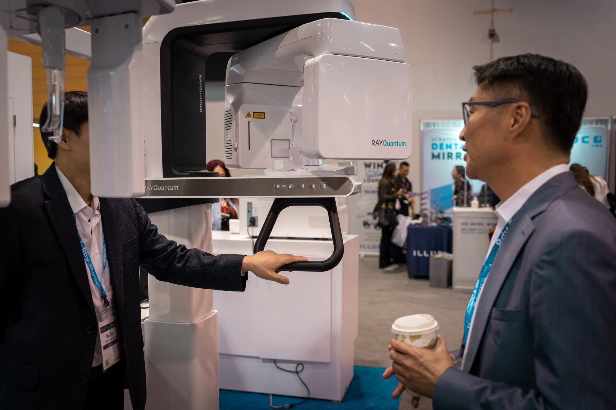 Two men at a dental conference examine a large piece of dental imaging equipment labeled 'RAYQuantum'. One man in a dark suit is operating the machine, while the other, also in a suit, holds a coffee cup and observes.