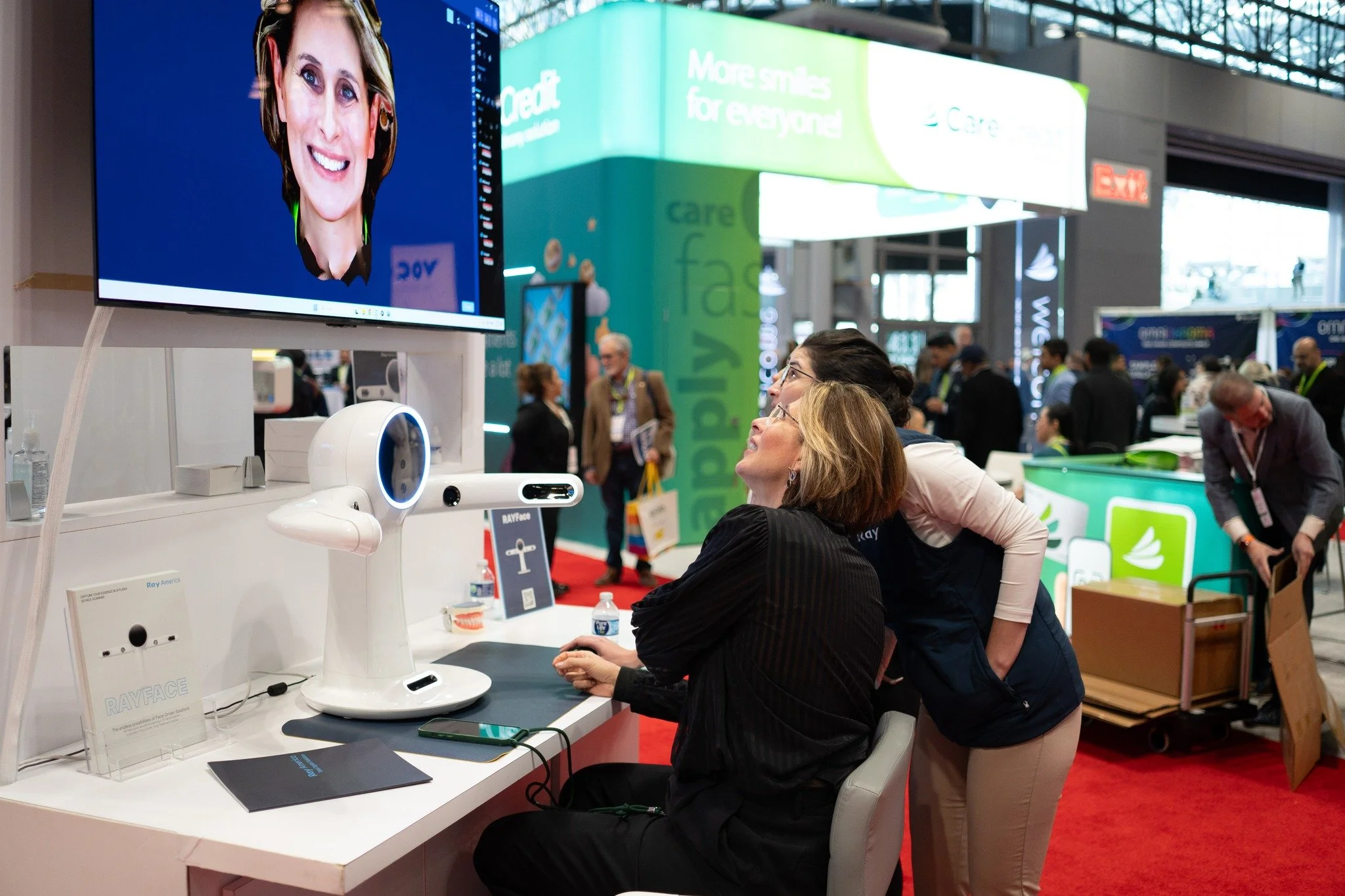 Two women at a trade show booth demonstrating RayFace technology with a large screen showing a smiling woman's face. Other attendees are in the background, with various booths and banners visible.