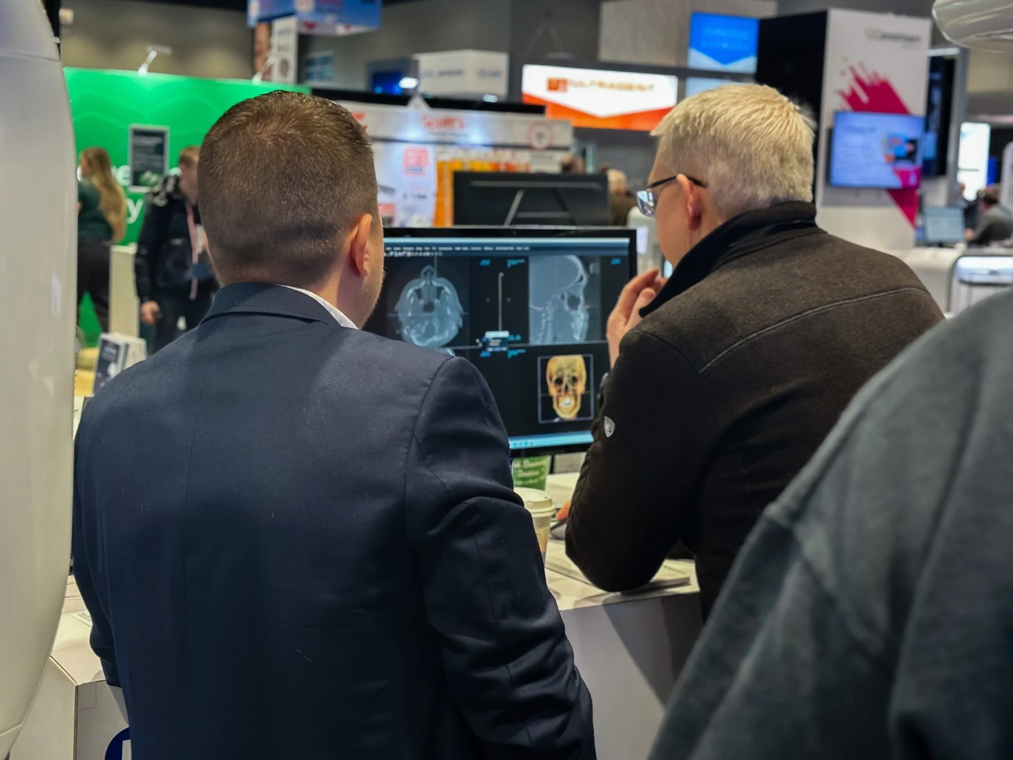 Two men are looking at medical scans of a skull and brain on a computer monitor at a busy trade show or conference.