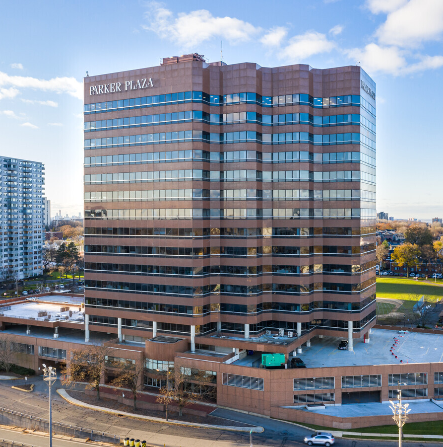 A tall office building with reflective glass windows labeled 'Parker Plaza' on the top left corner, situated in an urban area with other buildings, trees, and a street with cars in the foreground.