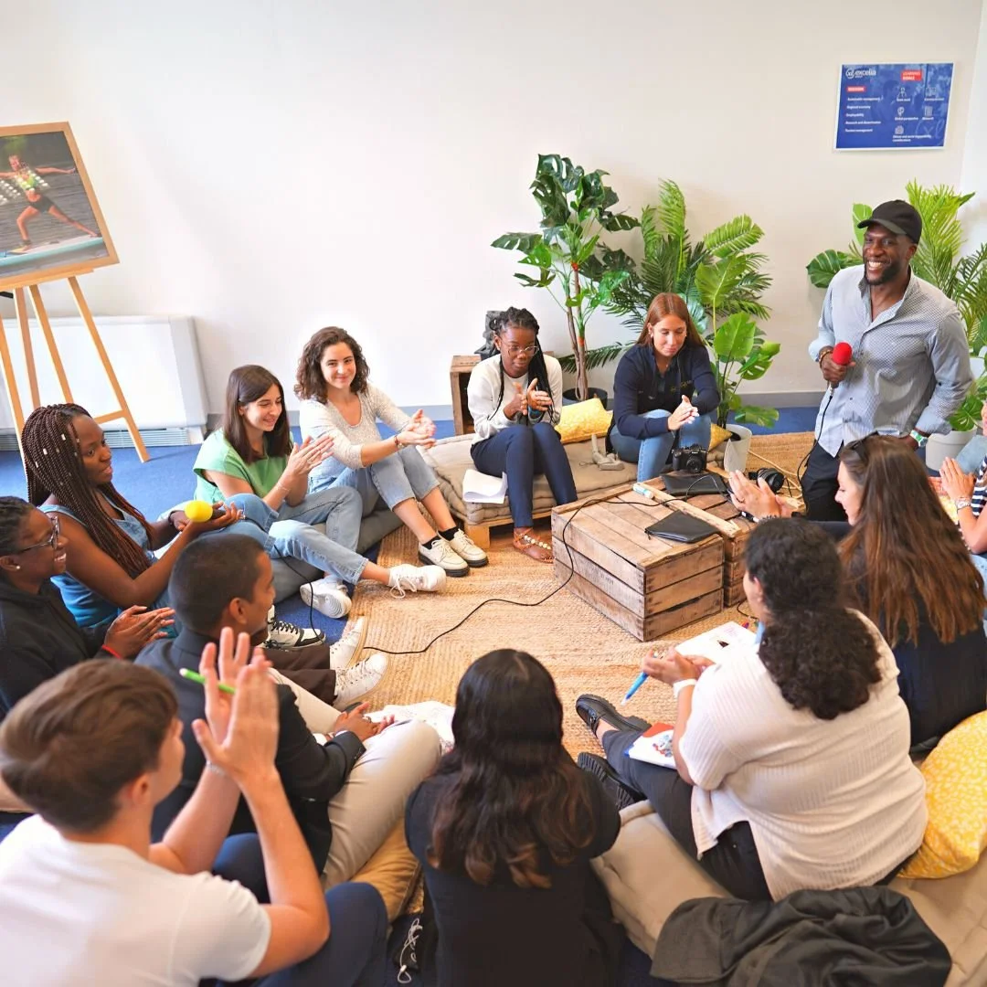 A group of people sitting on cushions and chairs in a circle, listening to a man standing with a microphone, in a room with plants and a white wall.