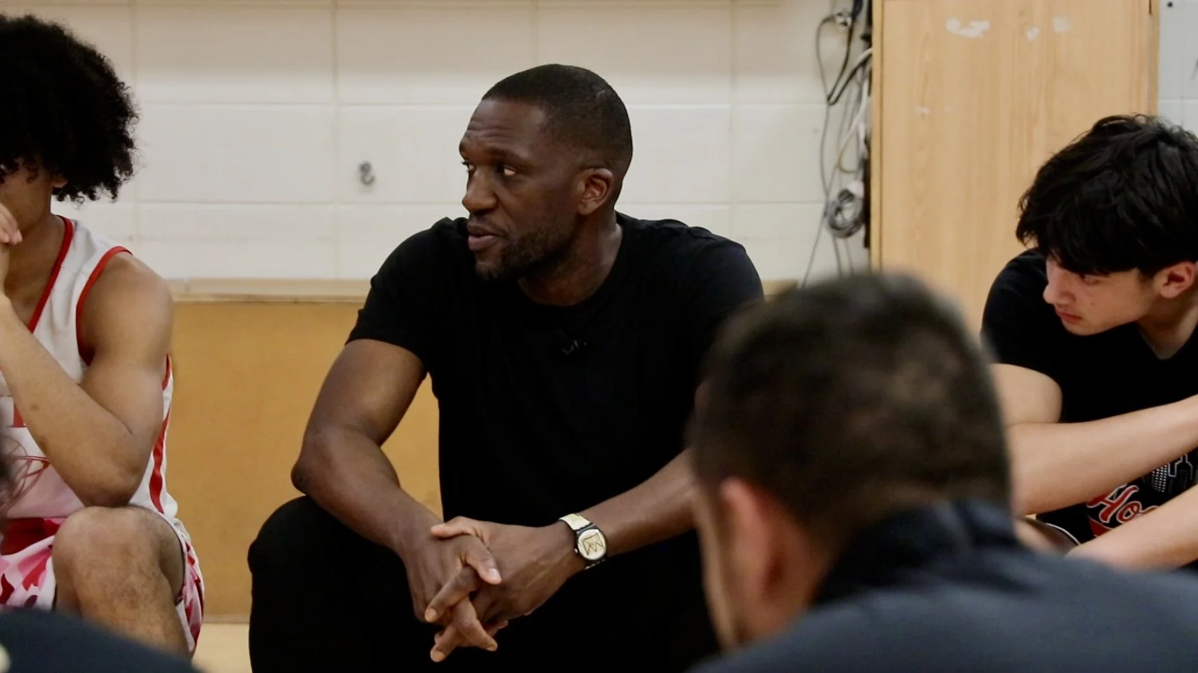 A man in a black shirt is sitting and speaking to a group of young people, some of whom are wearing sports jerseys, in an indoor setting with a wood-paneled wall and electrical cords in the background.