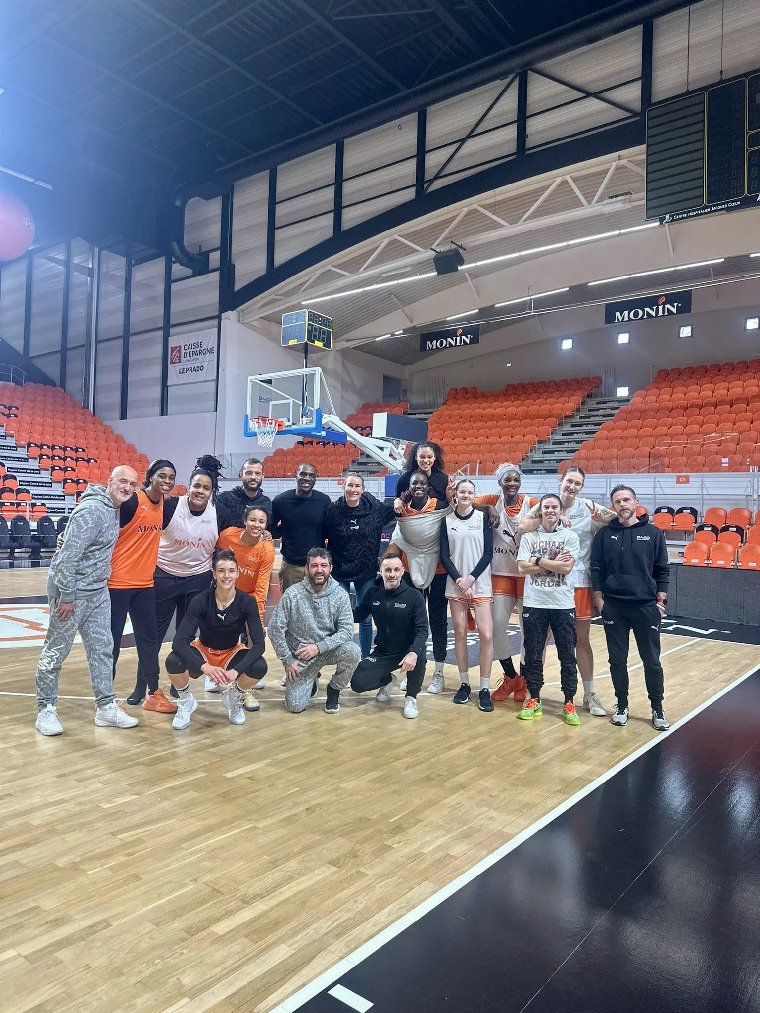 A group of basketball players and coaches posing for a photo on an indoor basketball court. The court has a wooden floor, orange seats in the background, and a basketball hoop. The players are in uniform, some in orange and others in white, and are smiling and making gestures.