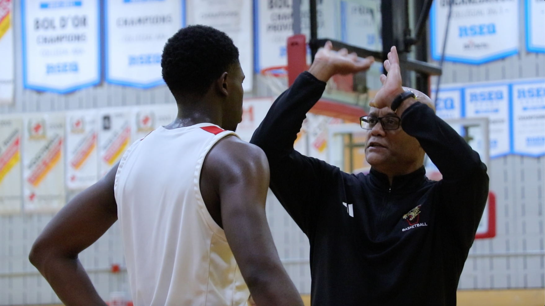 Basketball coach giving instructions to a player during practice in a gymnasium, with championship banners in the background.