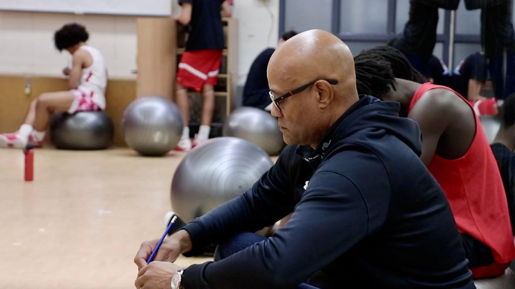 A man wearing glasses and a black jacket sitting on the floor with a pen in hand, surrounded by several exercise balls, in what appears to be a gym or sports facility. In the background, children are sitting and standing, wearing athletic attire.