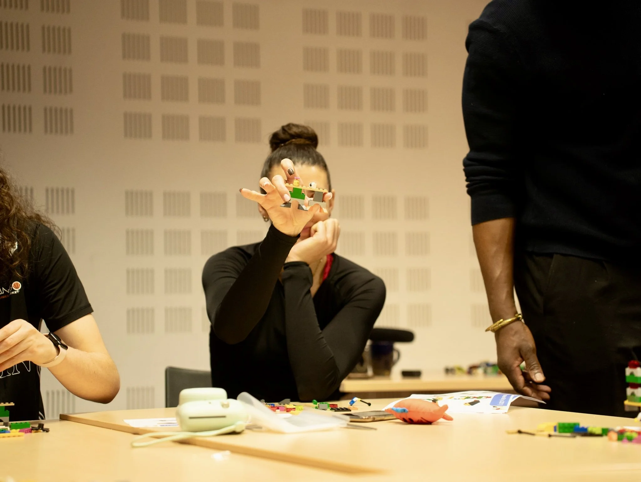 A woman is holding up a small Lego creation in a room with a beige wall and a patterned design. There are two other people partially visible, and various Lego pieces and tools are spread on the table in front of her.