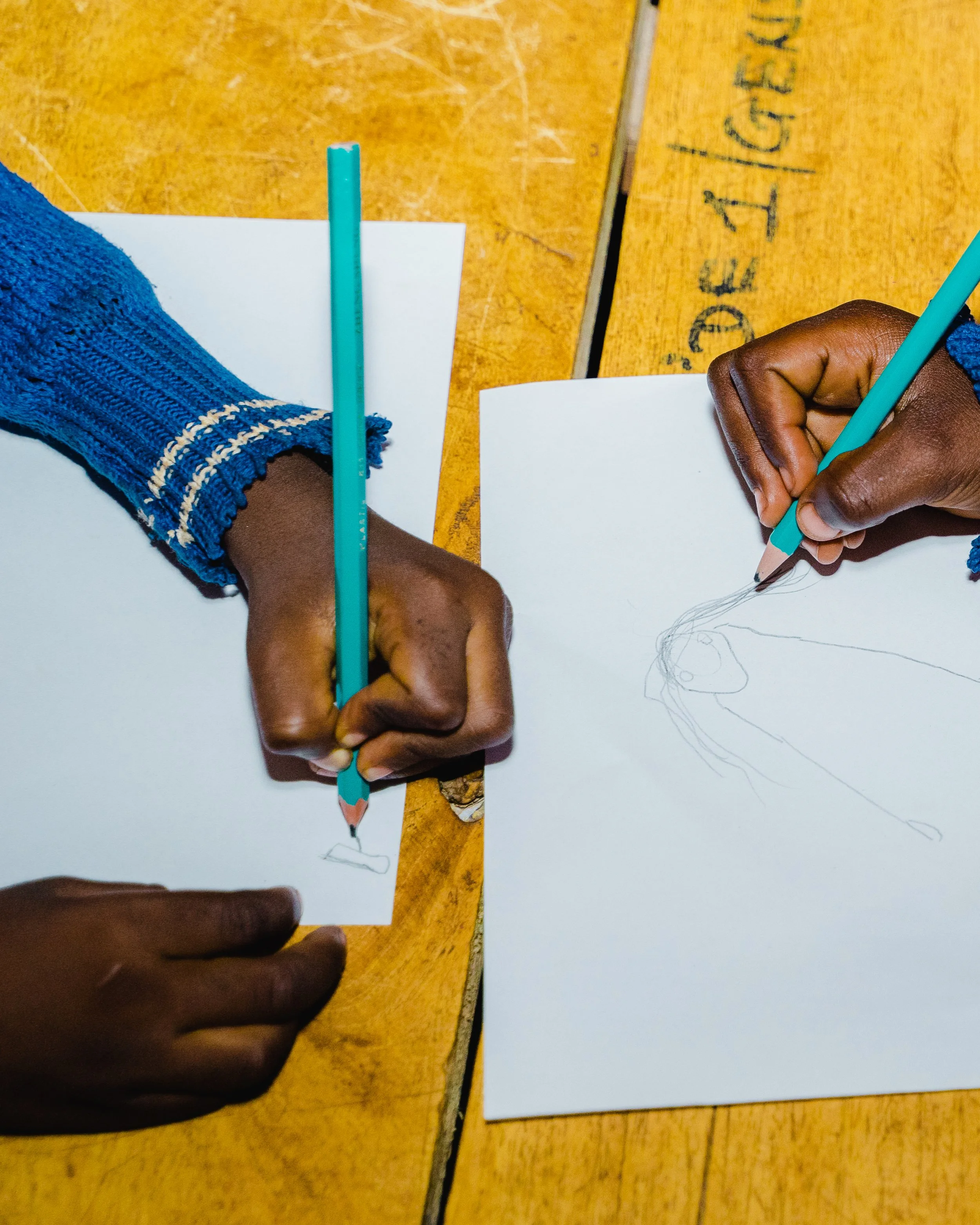 A person is drawing a face on a piece of paper using a turquoise colored pencil, with another hand resting on the paper, on a wooden table.
