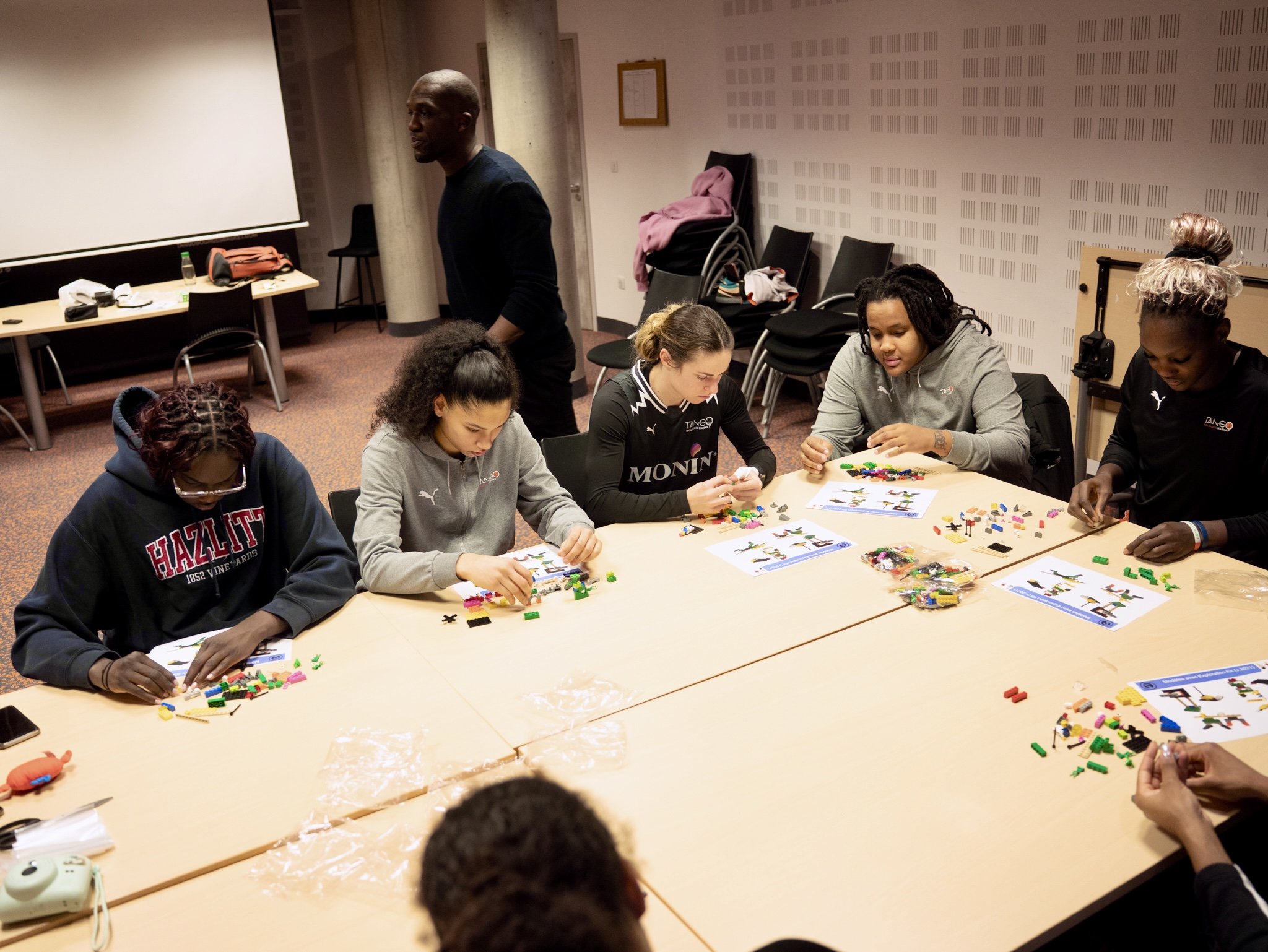 Group of diverse young women sitting at a table, assembling colorful LEGO pieces. An instructor or supervisor is standing nearby in a room with stacked chairs and a blank presentation screen.