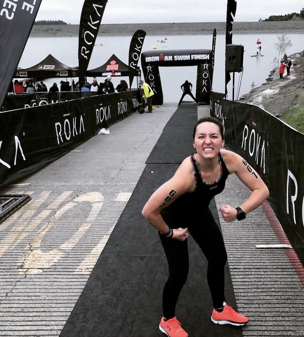 A woman in athletic gear showing a fierce expression with fists clenched at the finished line of an Ironman triathlon by a body of water. The event appears to be outdoors on a cloudy day, with spectators and a small boat visible in the background.