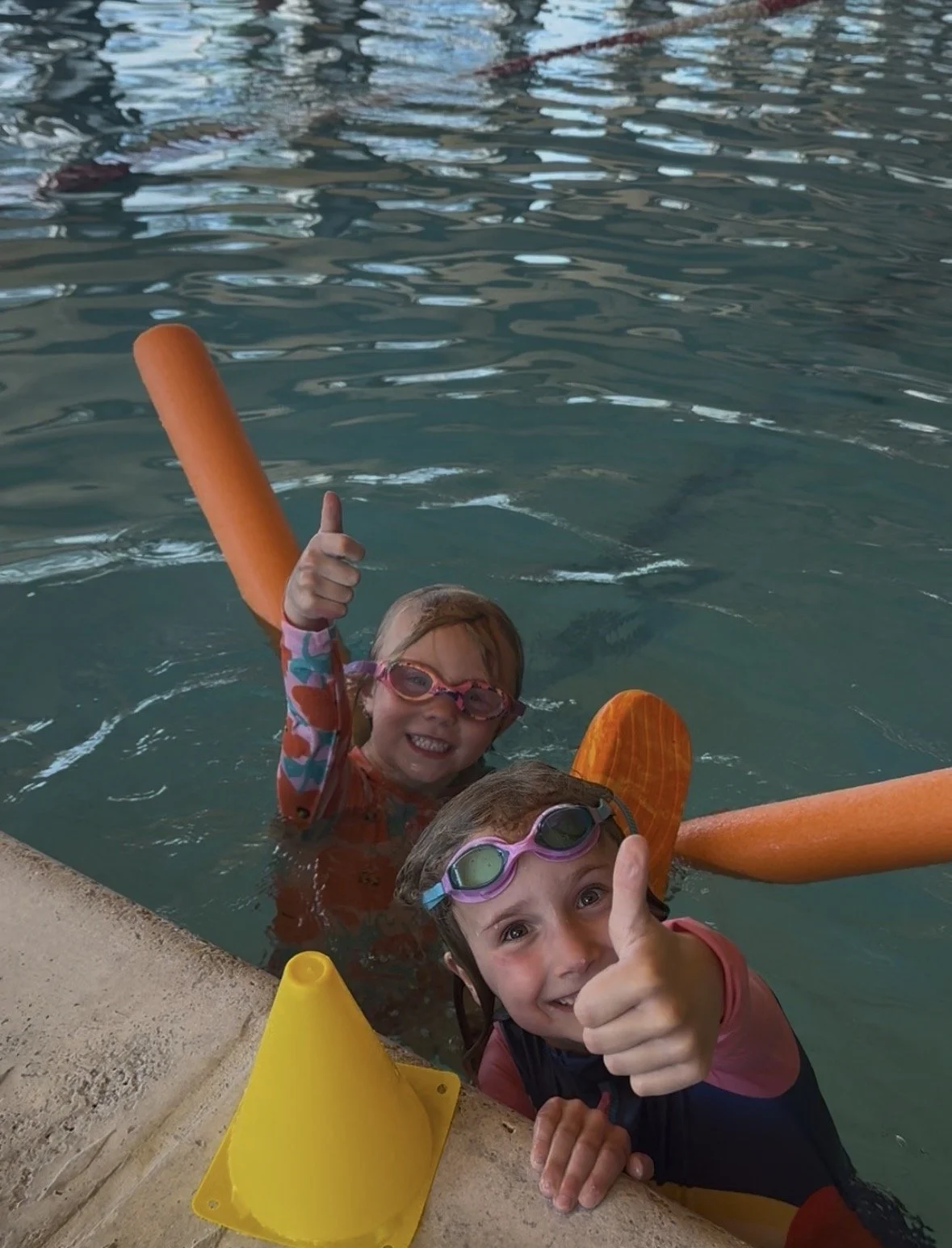 Two children in a swimming pool wearing goggles, holding orange foam noodles, smiling, giving thumbs up, near a yellow traffic cone.