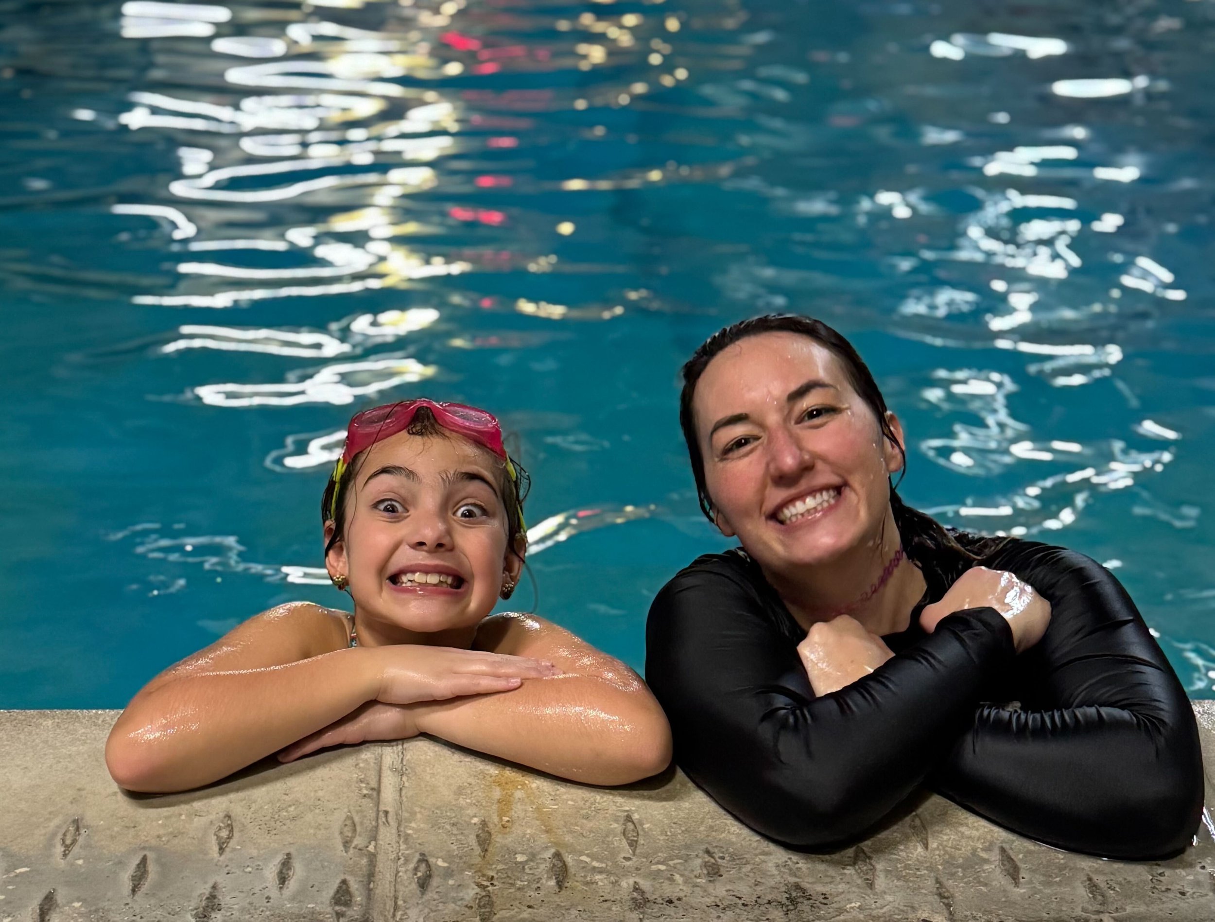 Two women and a girl smiling and leaning on the edge of a swimming pool, with water reflections above them.