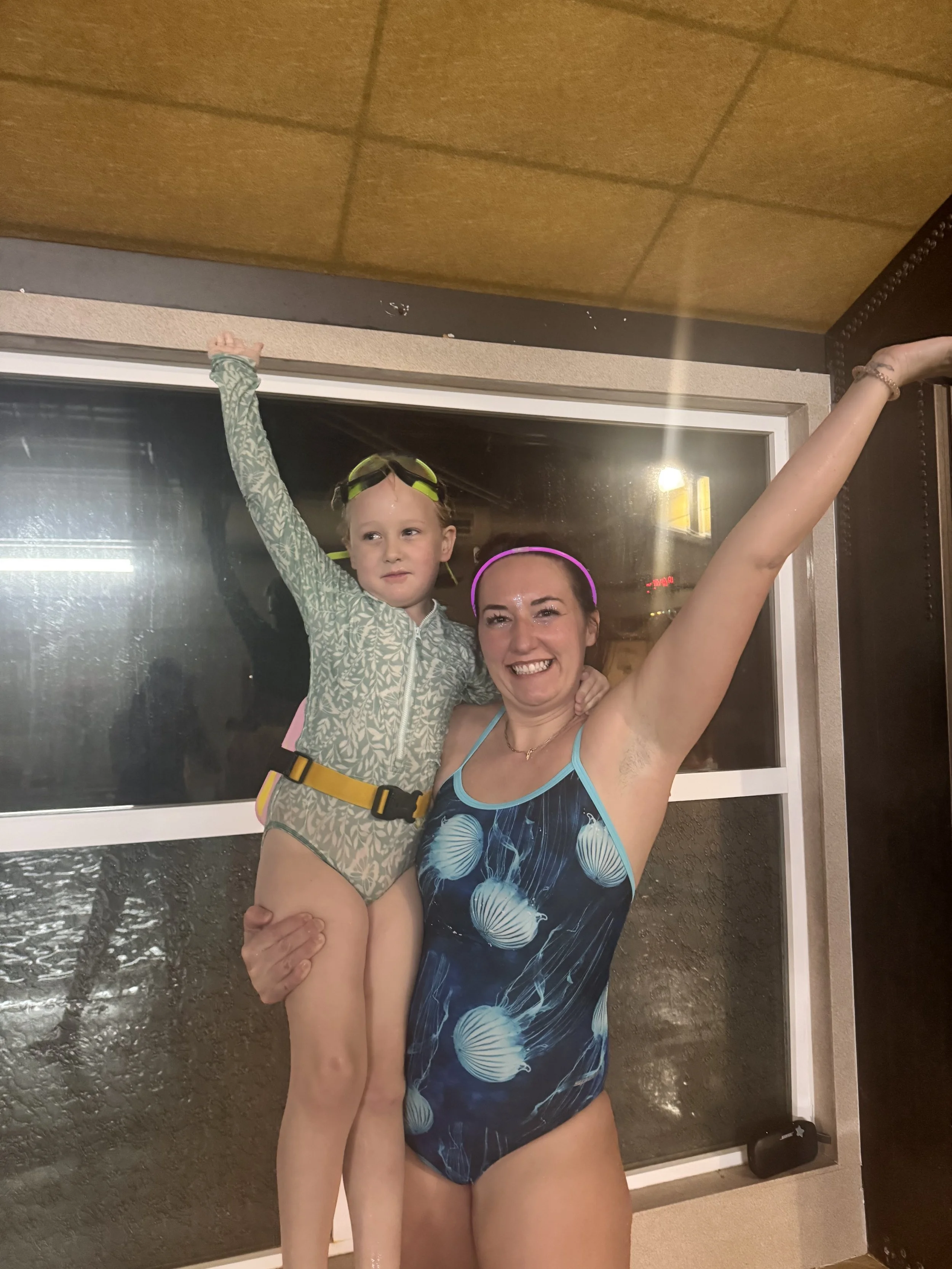 A woman is holding a young girl on her hip inside a building near a window with a dark exterior view. Both are smiling with arms raised, the woman wearing a swimsuit with jellyfish print and the girl wearing a patterned rash guard. The woman has a neon pink headband and a bracelet.