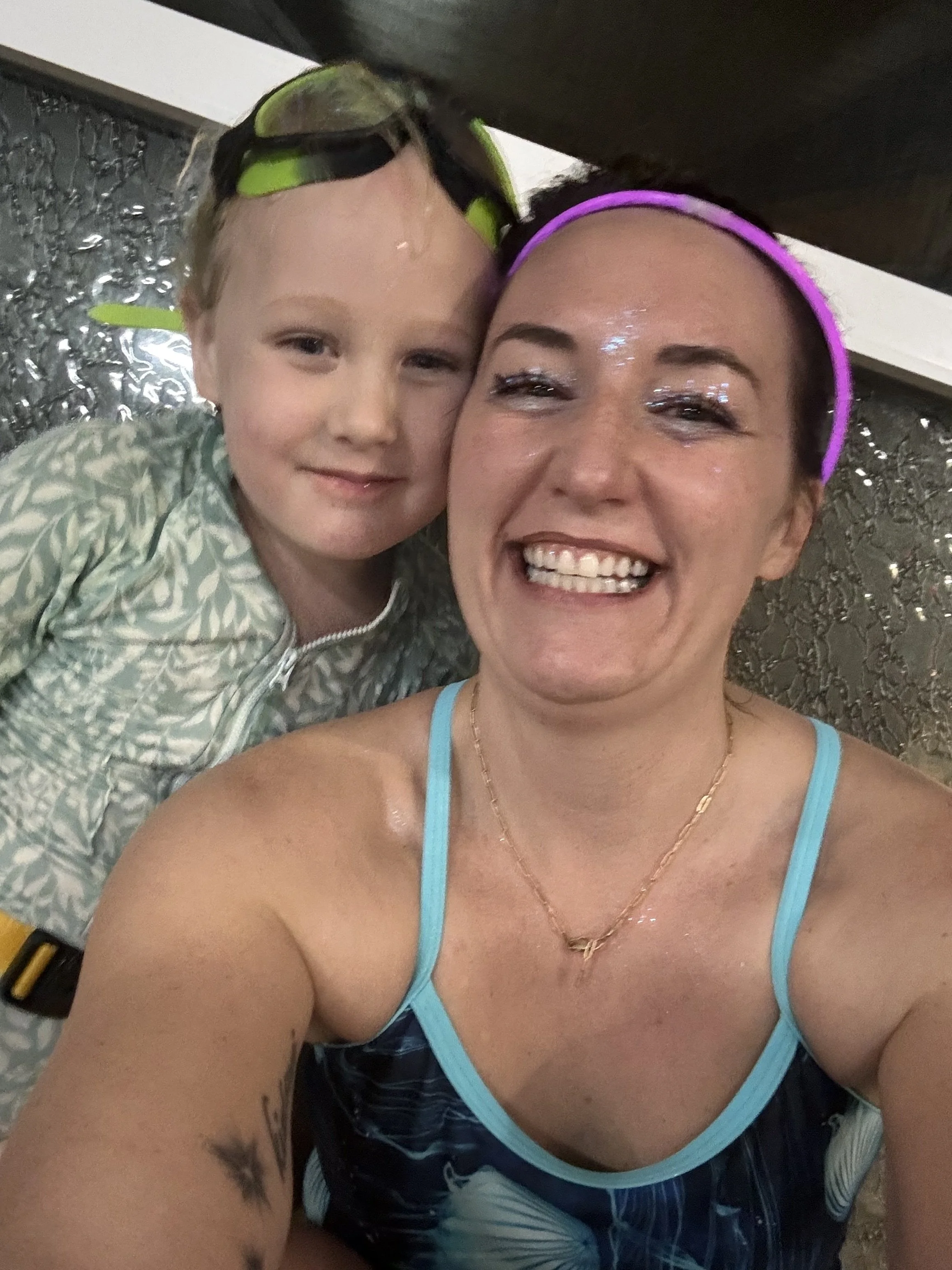 The woman coach is smiling broadly, wearing a purple headband, with wet hair and skin. The girl is wearing goggles  and a patterned suit. They are in an indoor pool area with a pool party, near a swimming pool.