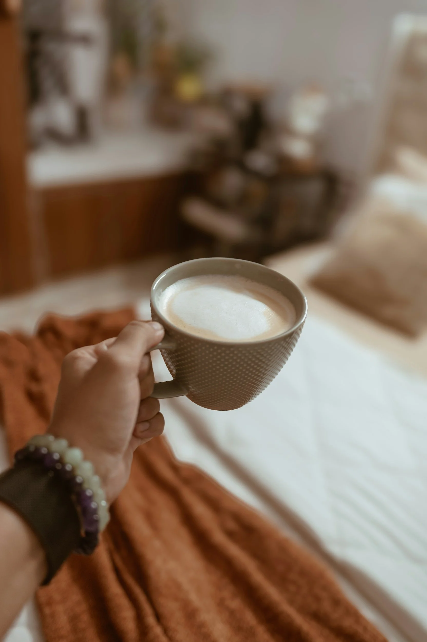 Person holding a beige mug with a white foam beverage, sitting on an orange blanket or fabric, with a blurred indoor background including a wooden surface and shelves.