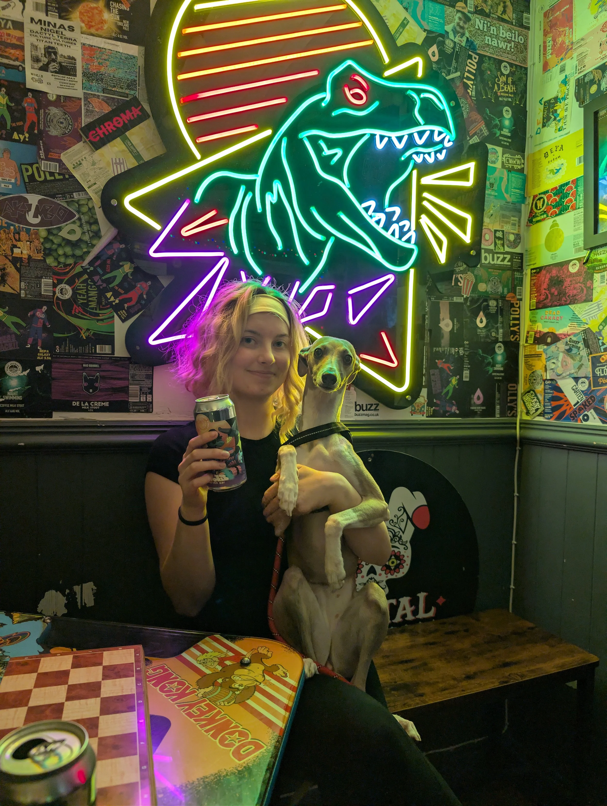 A person with curly blonde hair and a light-colored headband, holding a dog and a beverage, sitting at a table with a checkered board game in front of them inside a lively bar or arcade. Neon signs and colorful posters decorate the wall behind them.