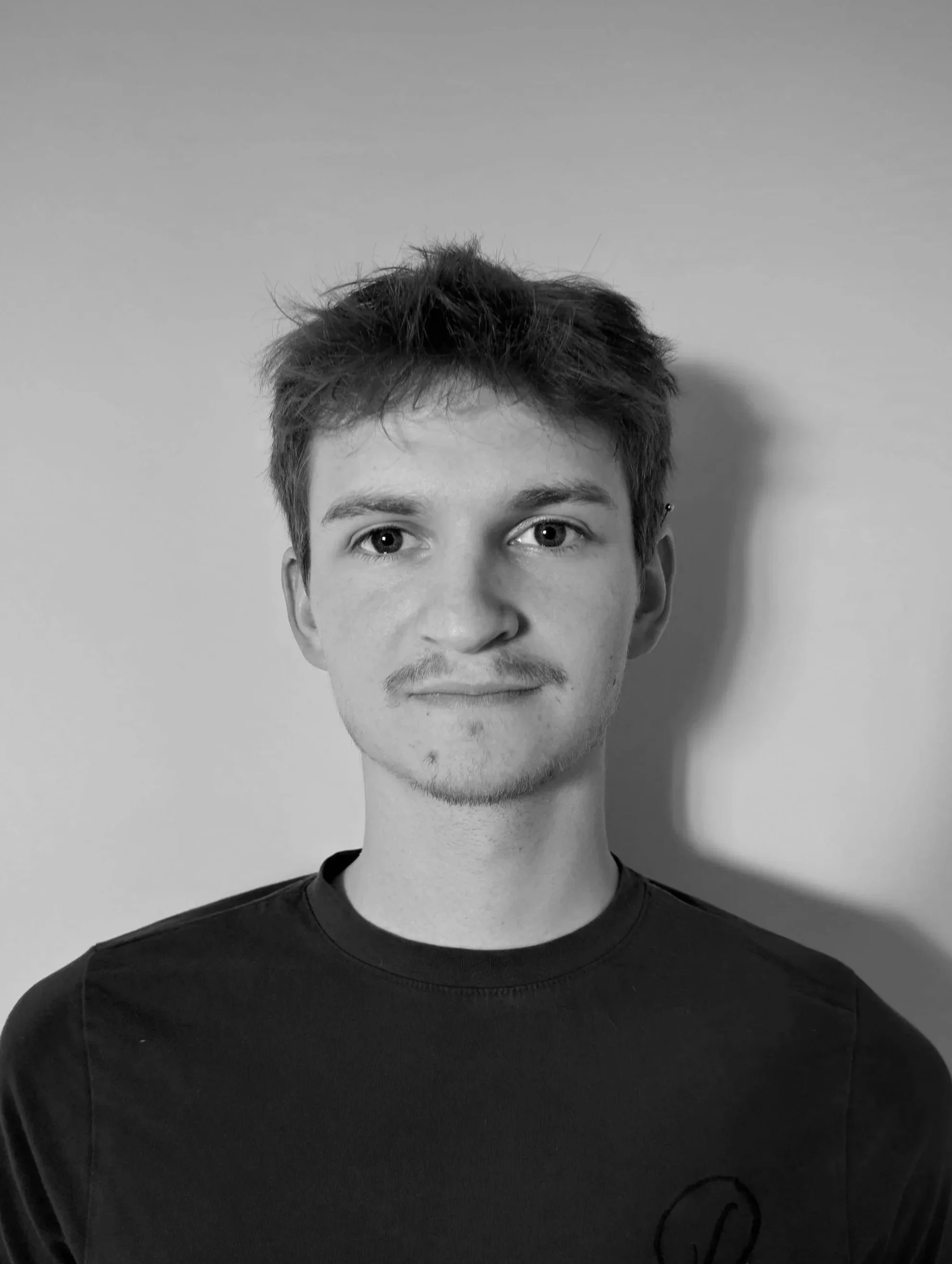 Black and white portrait of a young man with short, messy hair, looking directly into the camera, wearing a dark t-shirt, standing against a plain background.