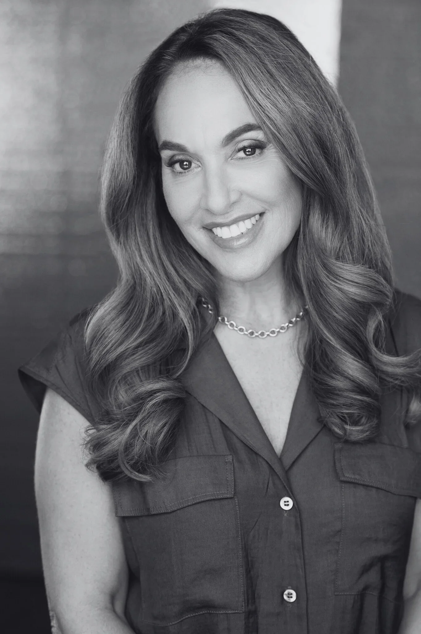 Black and white portrait of a woman with long wavy hair wearing a sleeveless button-up shirt with pockets, a chain necklace, and smiling.