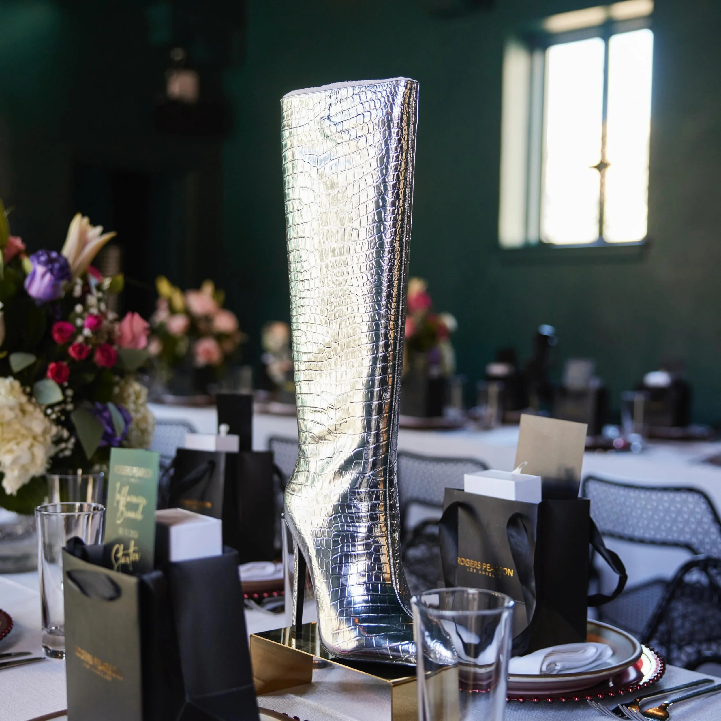 A shiny silver textured vase in a dining setting with floral arrangements and place settings on a table with a window in the background.
