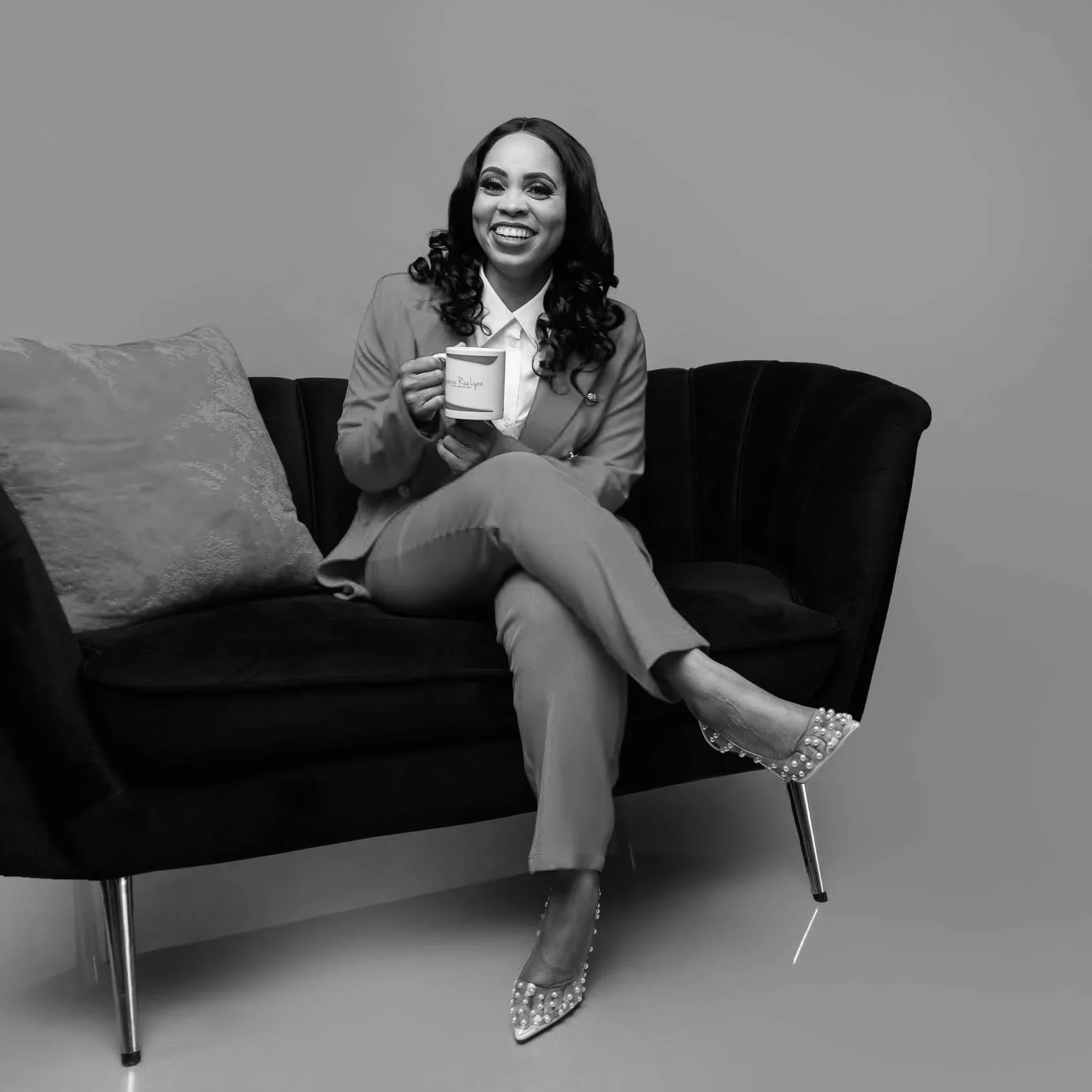 A woman with long wavy hair, smiling, sitting on a black sofa, holding a mug, wearing a light-colored blazer, patterned pants, and high-heeled shoes with pearl embellishments.