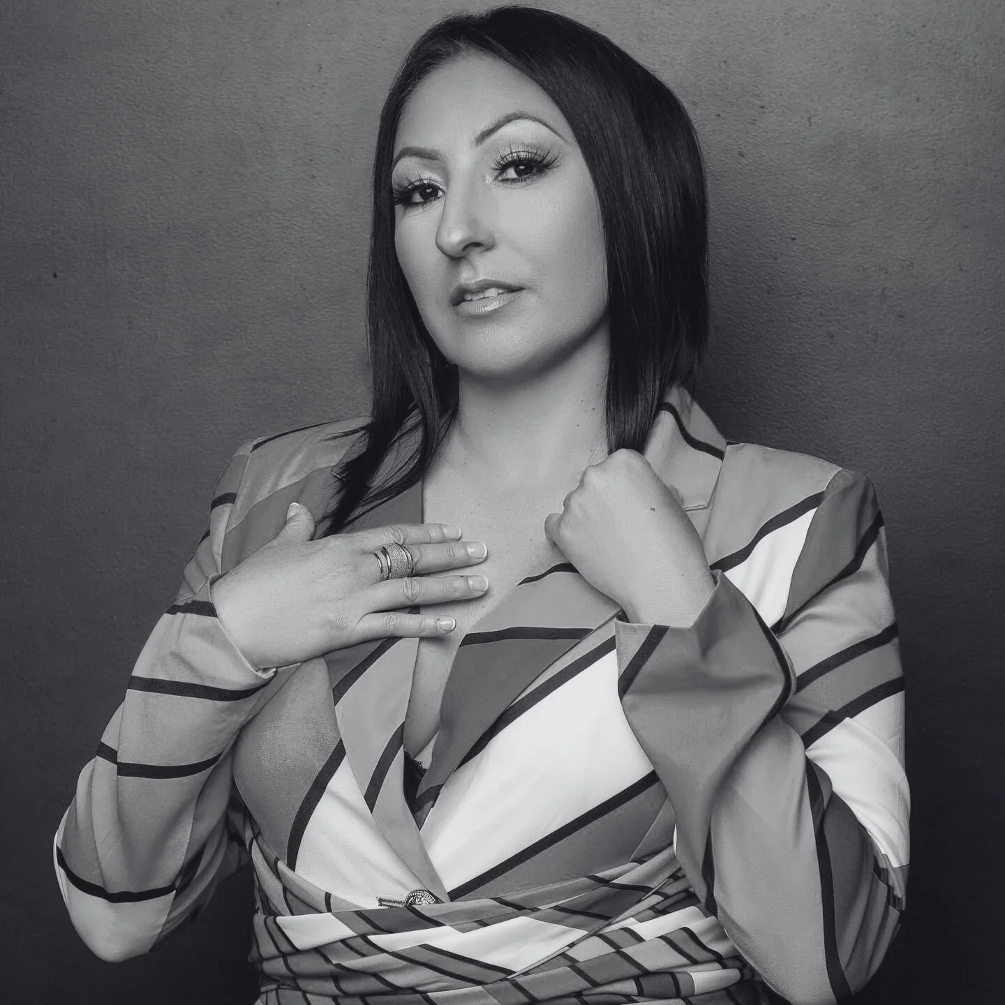 Black and white portrait of a woman with dark hair, wearing a striped shirt, looking at the camera with one hand on her chest and the other near her collar, against a plain background.