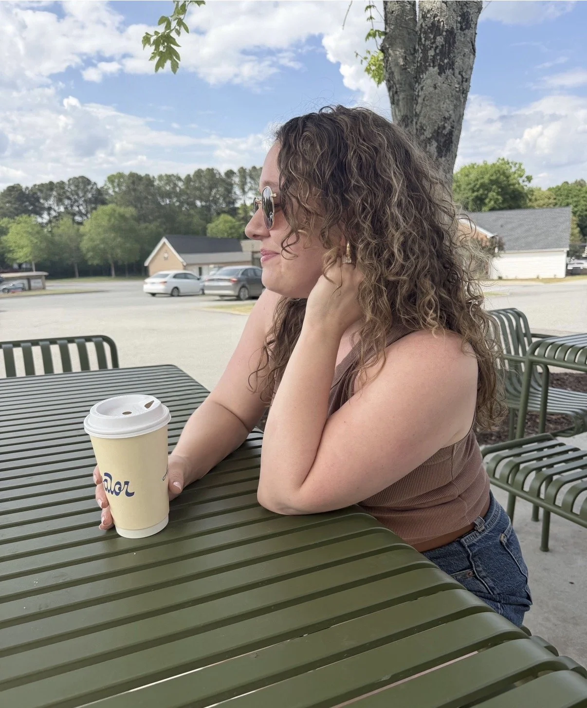 A woman with curly hair, wearing sunglasses, sitting at an outdoor table holding a coffee cup, looking to the side with trees, cars, and a cloudy sky in the background.