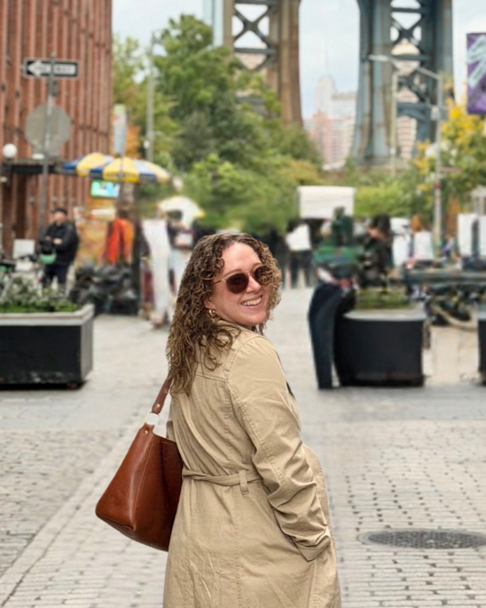 A woman with curly hair wearing sunglasses and a beige trench coat, smiling and looking over her shoulder on a city street with bridge structures in the background.